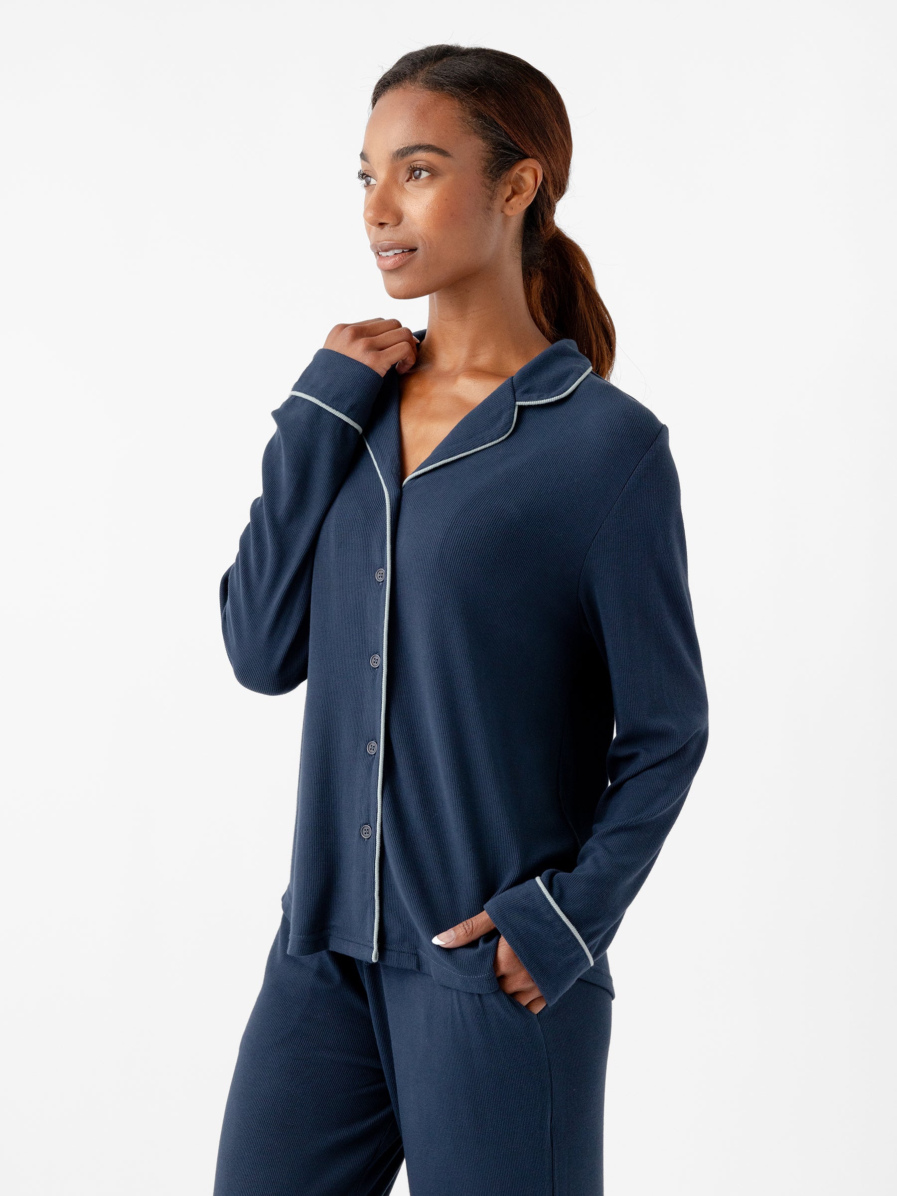 A woman with dark hair tied back is wearing Cozy Earth's Women's Bamboo Rib Knit Classic Long Sleeve Pajama Top in dark blue, featuring white piping along the collar and cuffs, paired with matching pajama pants. She is looking off to the side with a relaxed expression, against a plain white background. |Color:Eclipse