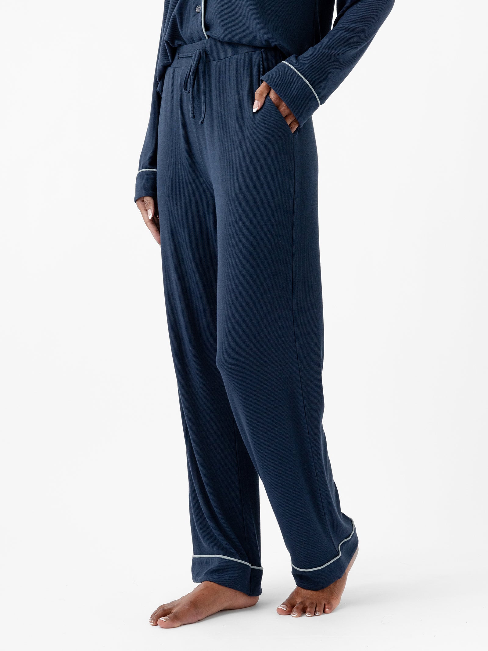 A person stands barefoot, wearing dark blue Women's Bamboo Rib Knit Classic Pajama Pant with a drawstring waist and white piping from Cozy Earth. The person has one hand in their pocket. The background is plain white. 