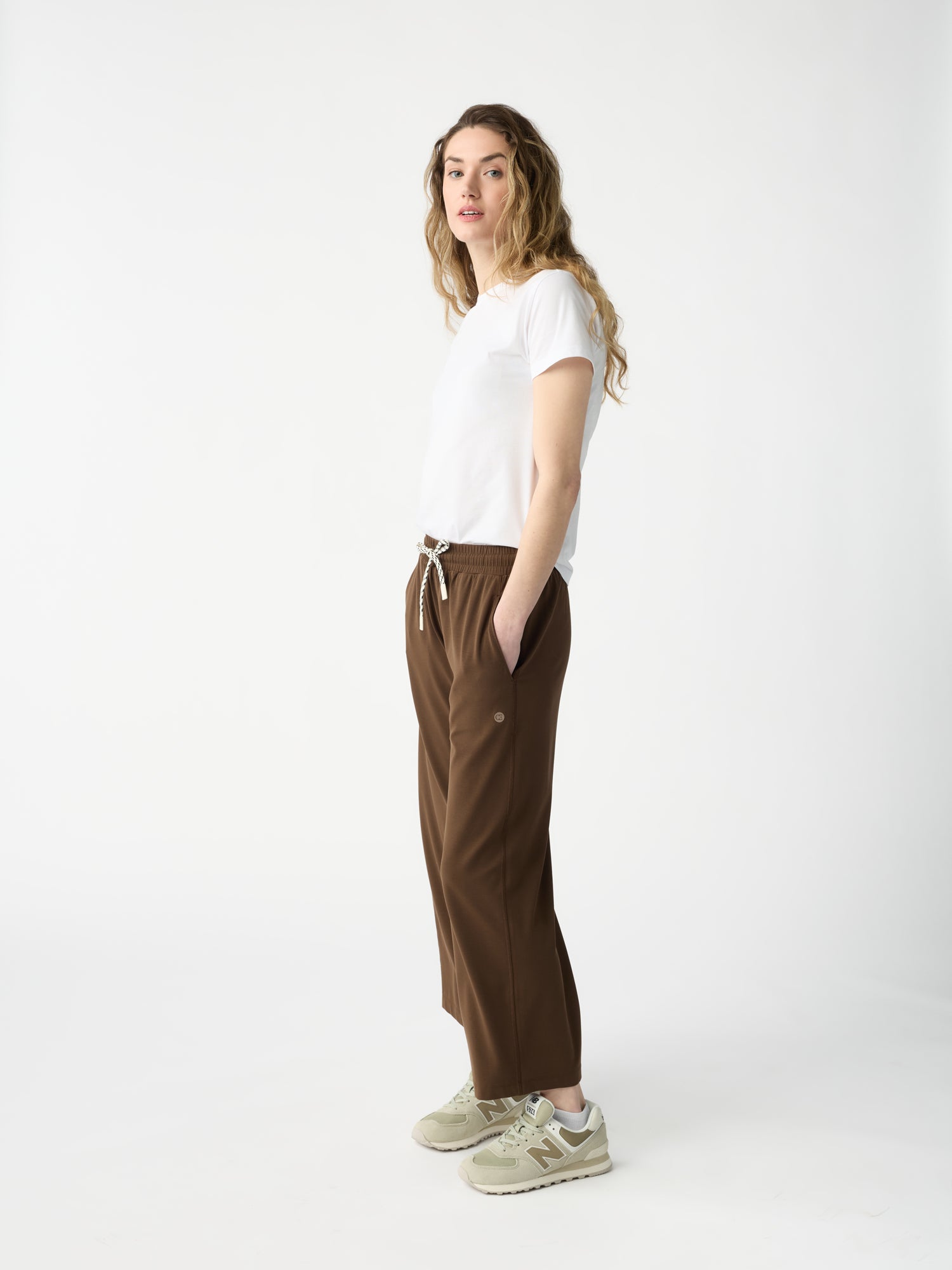 A person with long, wavy hair stands in profile against a white backdrop, wearing a white T-shirt, Cozy Earth's Women's Studio Cropped Wide Leg Pant in brown with a drawstring, and beige sneakers. Their hands are in pockets, and they have a neutral expression. 