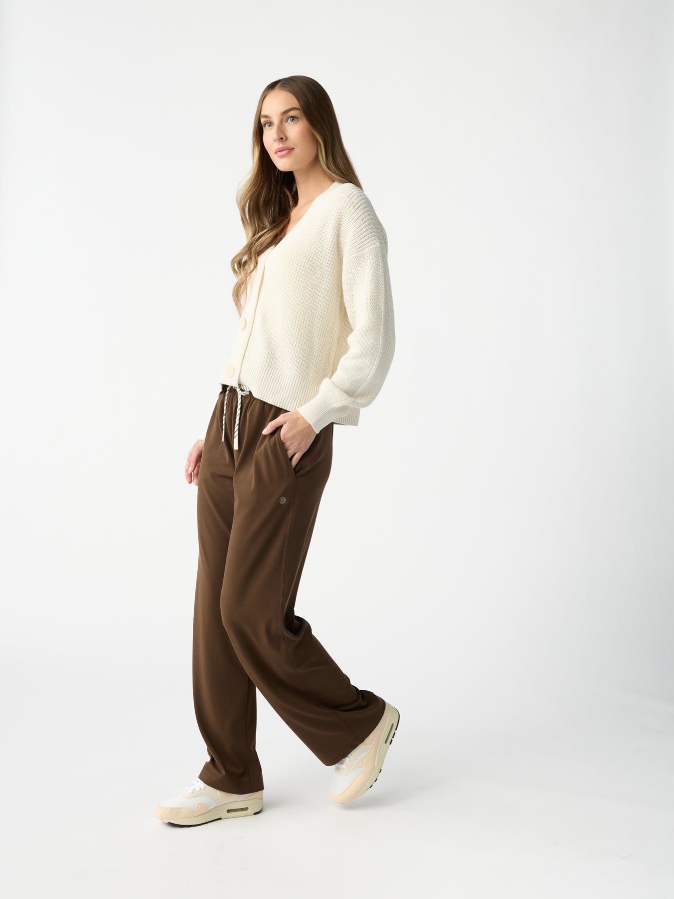 A person with long hair wears a cream-colored sweater, Cozy Earth's Women's Studio Wide Leg Pant in brown, and white sneakers against a white background, looking to the side with one foot slightly forward. |Color:Espresso