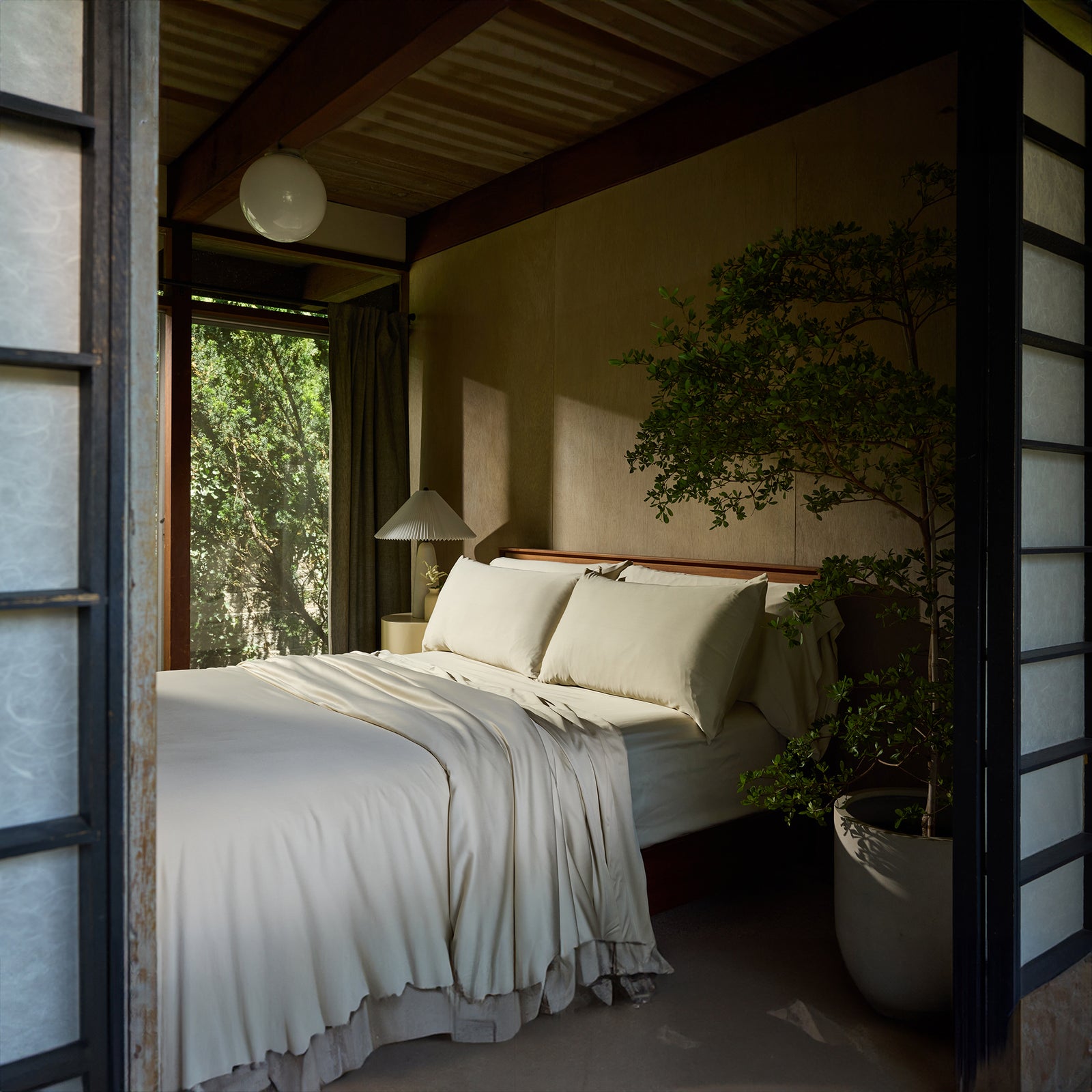 A cozy bedroom featuring a spacious bed adorned with a Bamboo Duvet Cover by Cozy Earth in cream, flanked by a bedside table with a lamp. A large potted plant rests at the foot of the bed. The room is framed by wooden panels and shoji screens, with green foliage visible through an open window. 