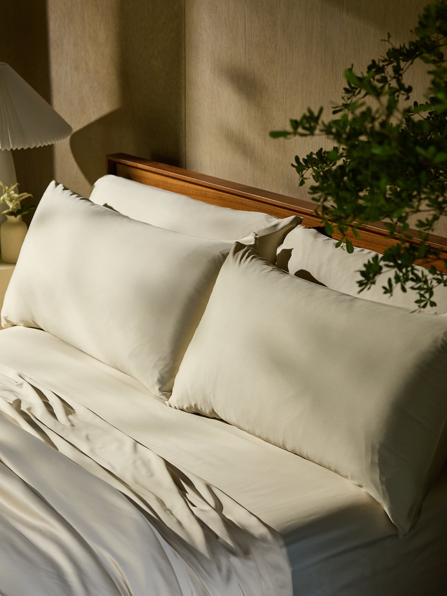 A neatly made bed with Cozy Earth's Bamboo Fitted sheet and two large white pillows is positioned against a wooden headboard. To the side, a small lamp with a white shade sits on a nightstand. Green foliage partially frames the right side of the image, adding a touch of nature. 