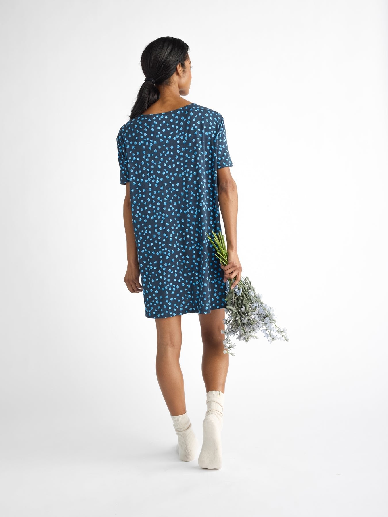 A long-haired person in a blue Women's Bamboo Stretch-Knit Sleep Dress from Cozy Earth and white socks holds a small flower bouquet in their right hand, walking away against a plain white backdrop. 