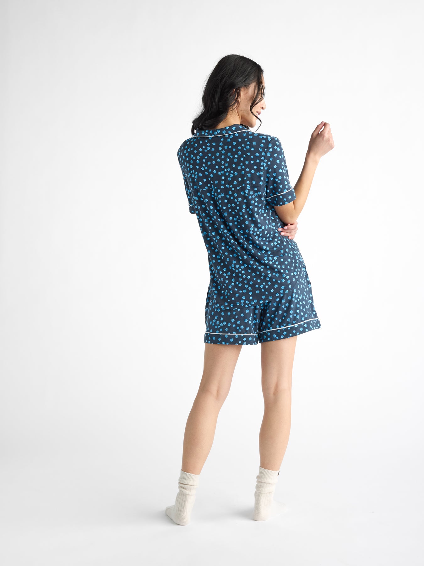 A person with long hair stands playfully facing away in Cozy Earth's Women's Bamboo Stretch-Knit Short Sleeve Pajama Set. They wear light socks against a plain, well-lit background. 