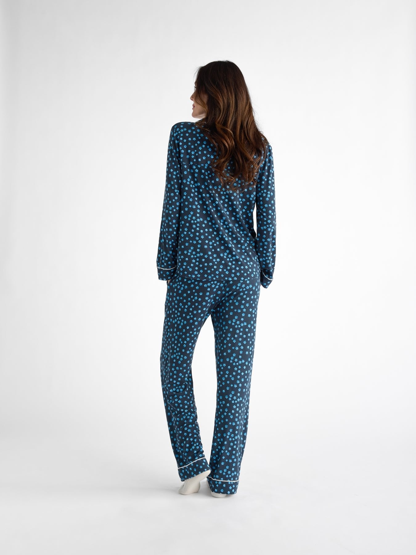 A person with long brown hair is standing and facing away against a plain white background, wearing the Women's Bamboo Stretch-Knit Long Sleeve Pajama Set from Cozy Earth in blue with a white polka dot pattern. 