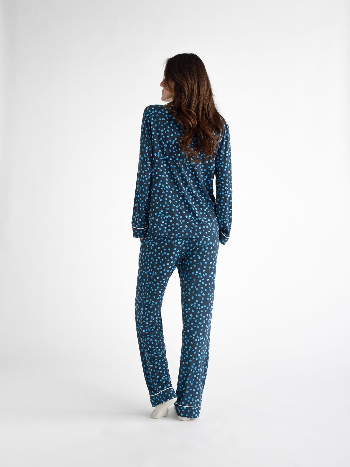 A person, back to the camera, wears Cozy Earth Women's Bamboo Stretch-Knit Long Sleeve Pajama Top in blue with white polka dots. They have long brown hair and are barefoot against a plain white background. 