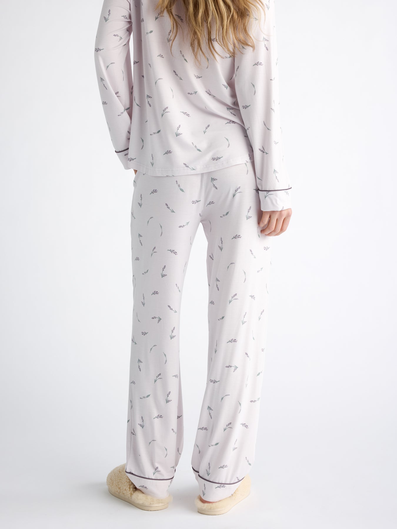 A person with long hair wears the Women's Bamboo Stretch-Knit Long Sleeve Pajama Set by Cozy Earth, featuring small, delicate patterns. They stand back turned with hands by their sides wearing fluffy slippers, set against a plain white background. 