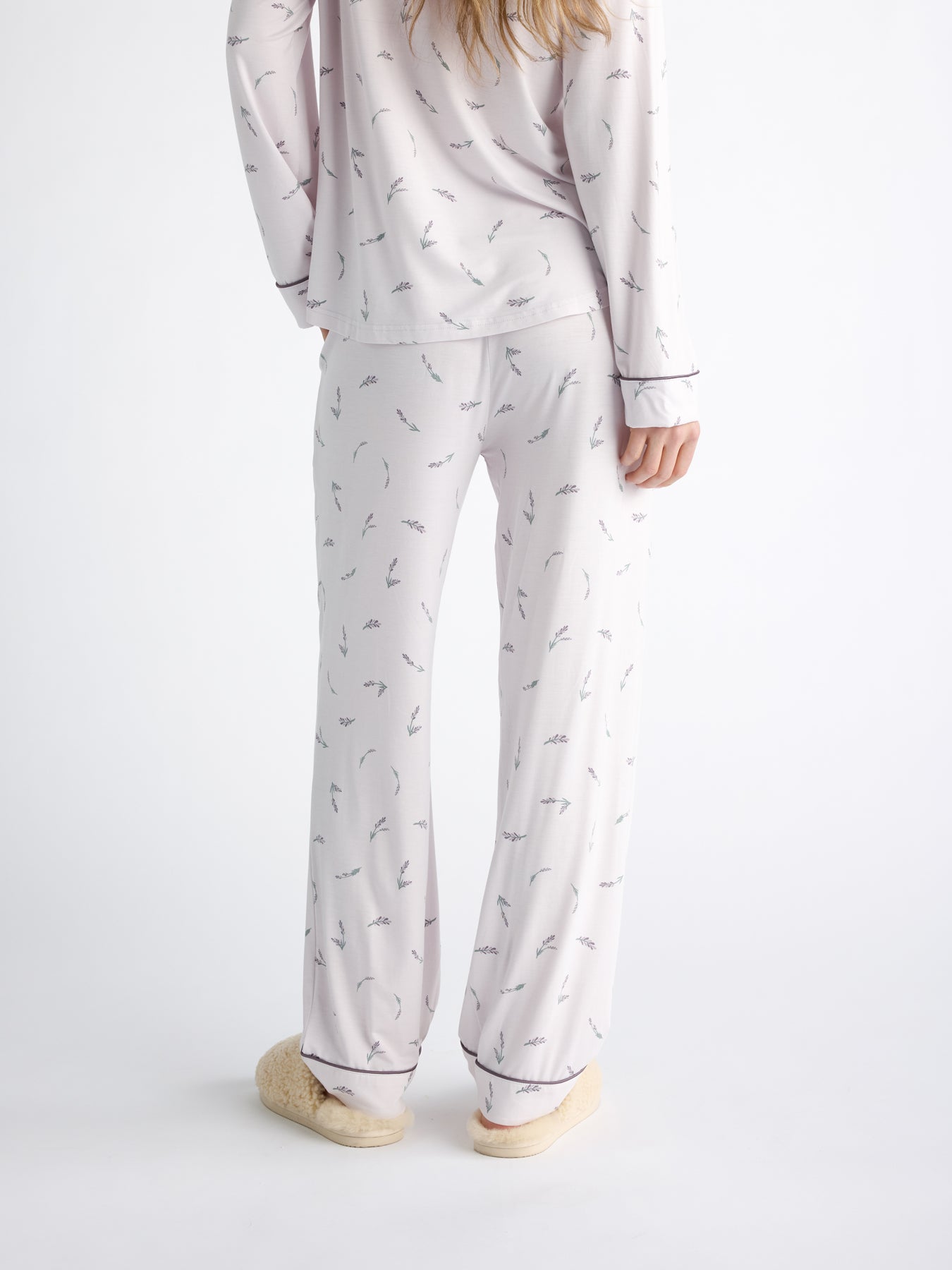 Back view of someone wearing Cozy Earth Women's Bamboo Stretch-Knit Pajama Pant in a light color with a subtle pattern, paired with fluffy slippers. The person has long hair and stands against a plain white background. 