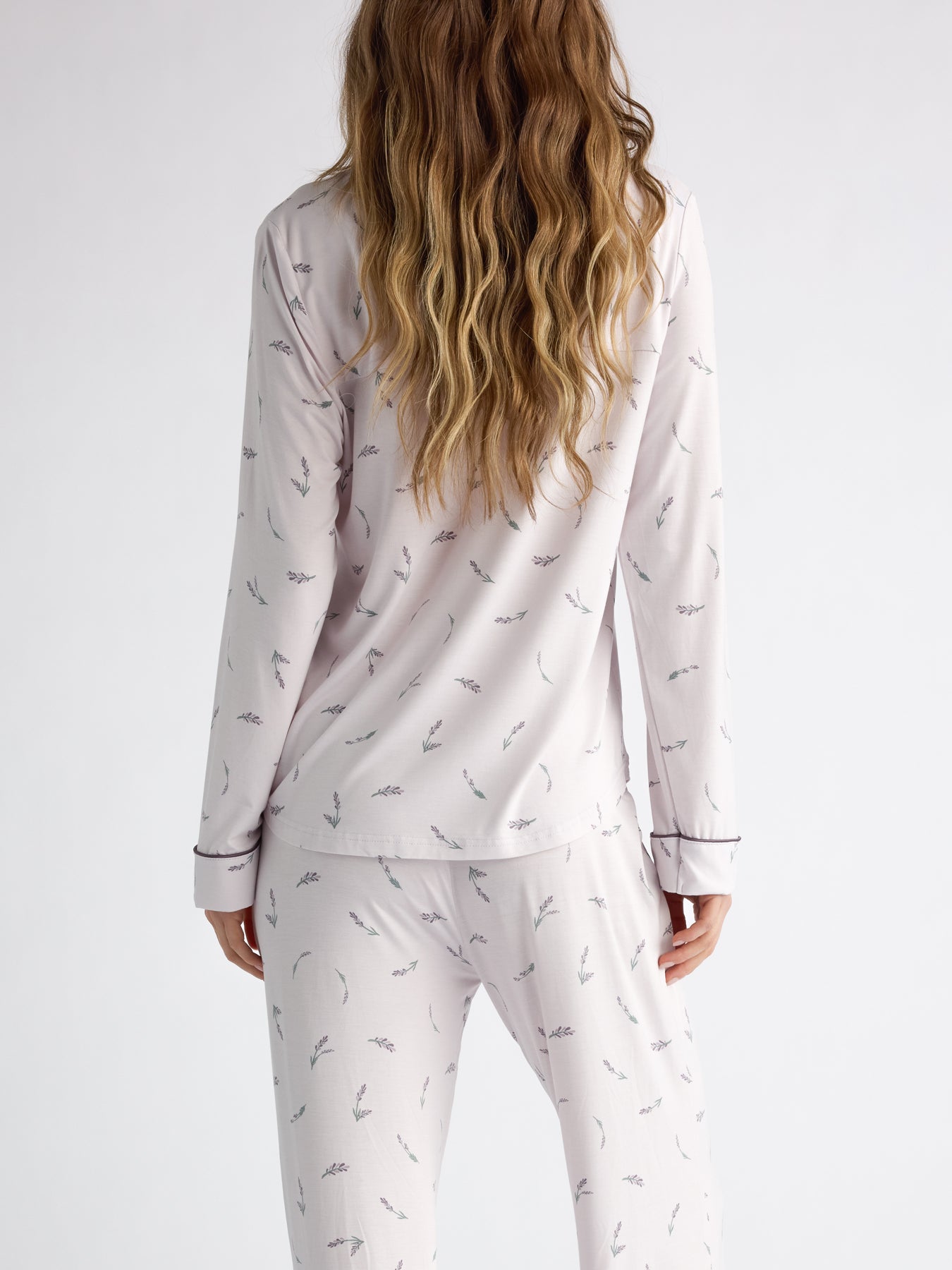 A person with long wavy hair faces away, wearing Cozy Earth Women's Bamboo Stretch-Knit Long Sleeve Pajama Top in a light color featuring a subtle pattern against a plain white background. 