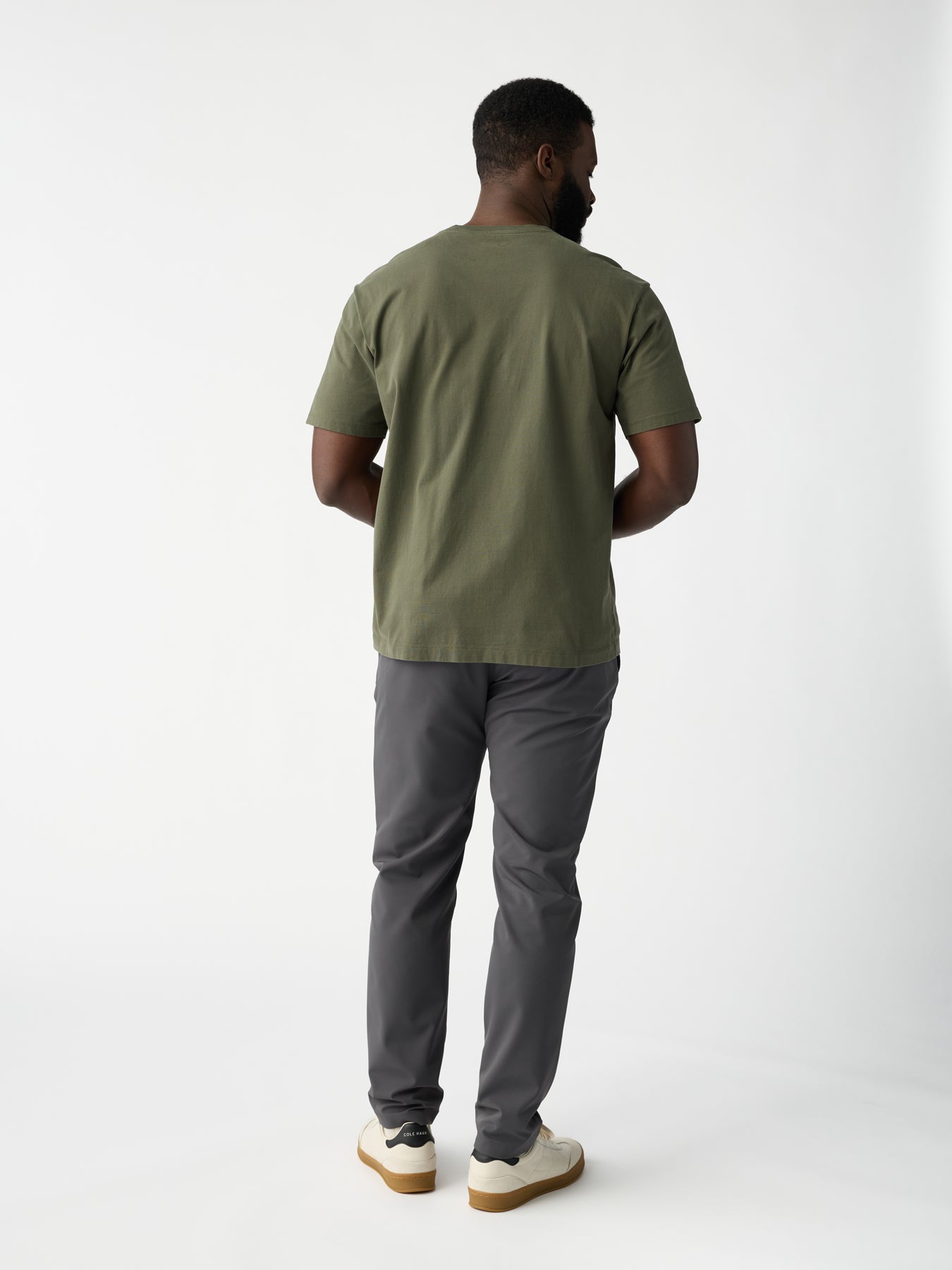 A person stands with their back turned, wearing a plain green T-shirt, Men's Everywhere Pant from Cozy Earth in gray, and white sneakers with brown soles against a plain white background. 