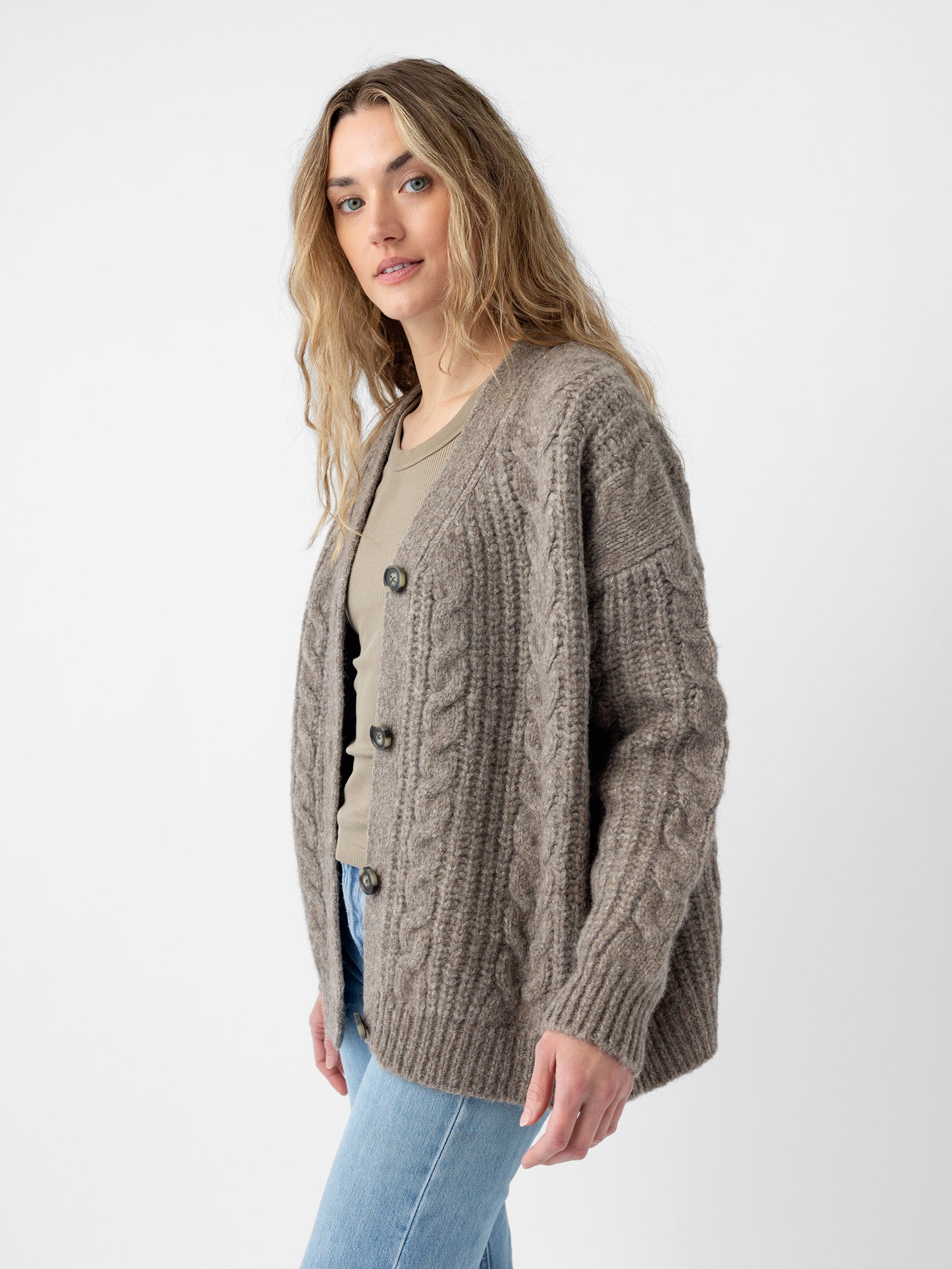 A person with long hair is wearing the "Oversized Cable Knit Cardigan" by Cozy Earth, along with a light shirt. They are standing against a plain background, also dressed in light blue jeans, and gazing toward the camera. 