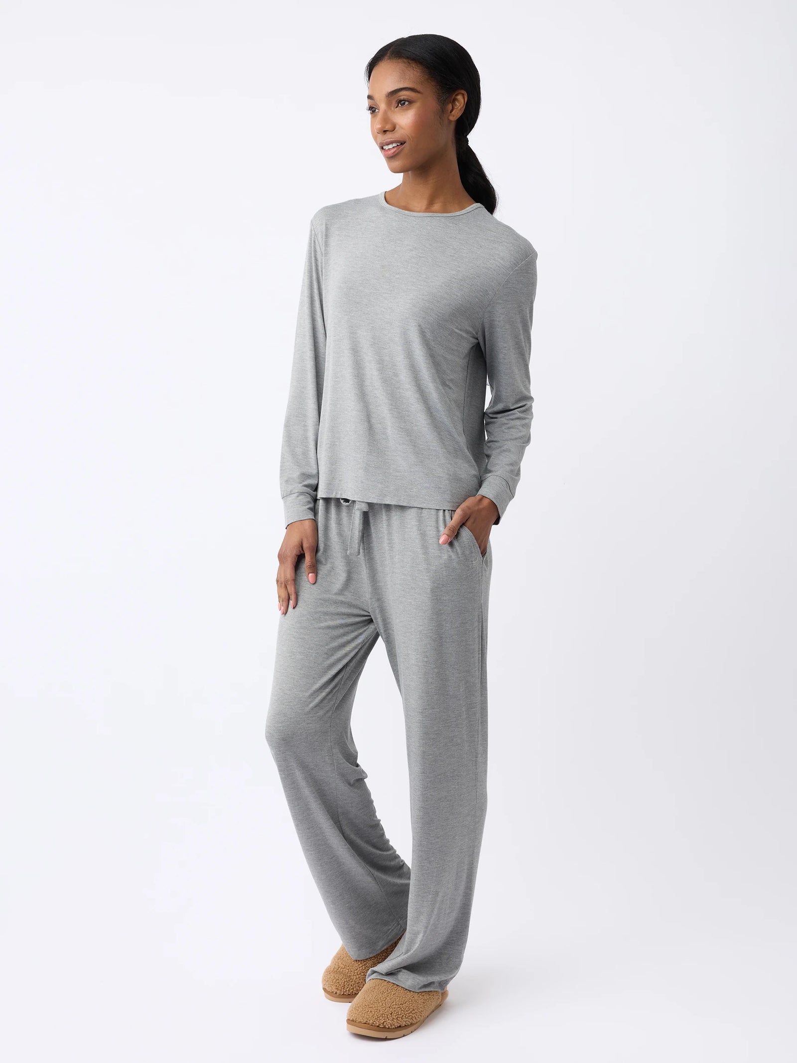 A woman wearing the Women's Stretch Knit Bamboo Long Sleeve Lounge Tee from Cozy Earth stands against a plain white background in a gray loungewear set, featuring a long-sleeve top and matching pants. With one hand in their pocket and brown slippers on, they are smiling slightly. 