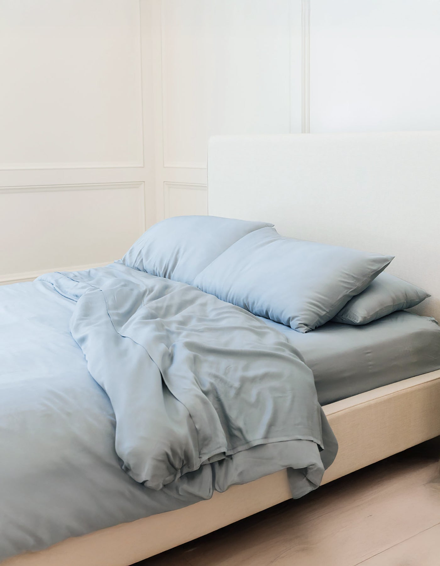 A neatly made bed features the Bamboo Sheet Set by Cozy Earth in light blue, with pillows against a white paneled wall. The bed has a cream-colored upholstered headboard, creating a light and airy atmosphere.