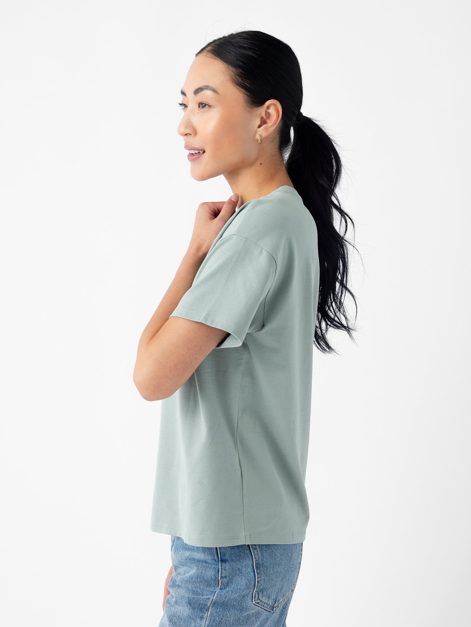 A person with long black hair tied back in a ponytail is standing sideways, looking to the left. They are wearing Cozy Earth's Women's All Day Tee in light green and blue jeans against a plain white background. 