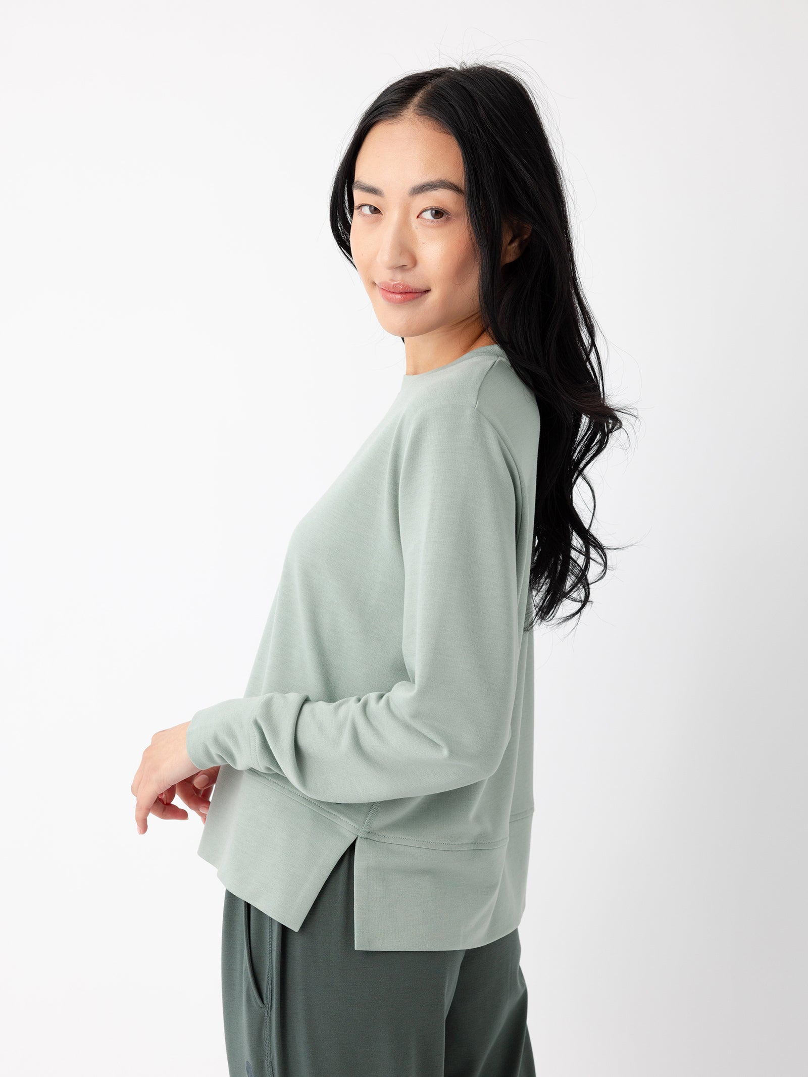 A person with long dark hair is standing against a white background, wearing a light green Women's StudioLite Crewneck from Cozy Earth and dark green pants. The person is smiling and looking over their shoulder. 