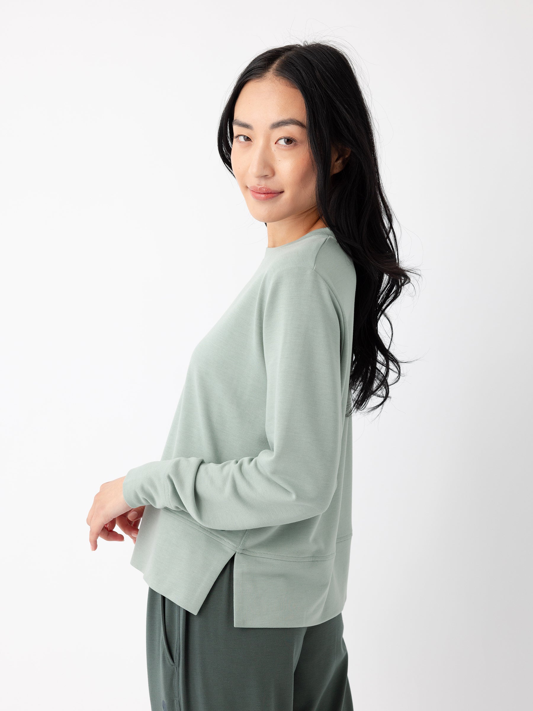 A person with long dark hair is standing against a white background, wearing a light green Women's StudioLite Crewneck from Cozy Earth and dark green pants. The person is smiling and looking over their shoulder. |Color:Haze