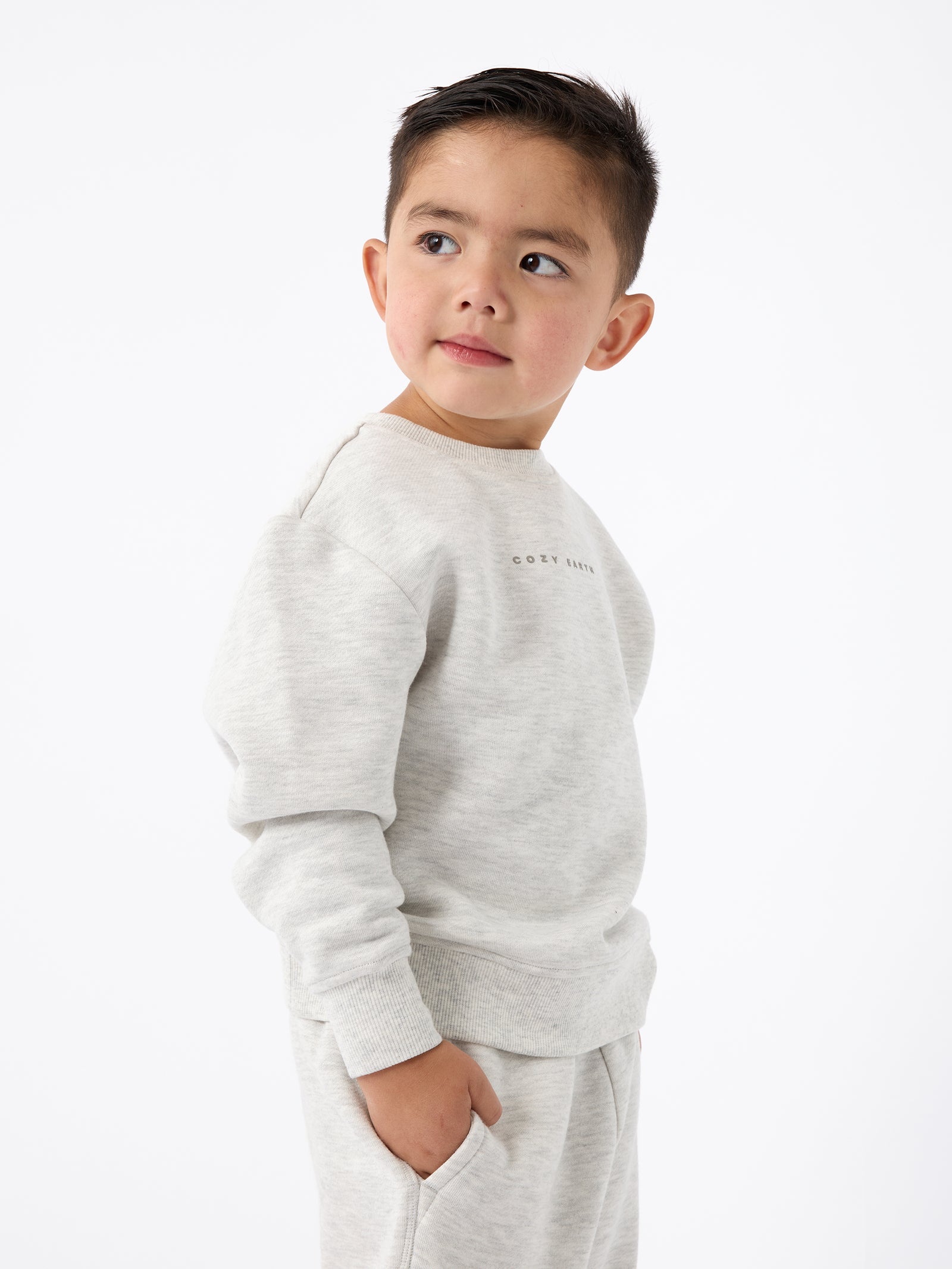 A young child with short hair is wearing Cozy Earth's light gray Kid's CityScape Crewneck sweatshirt and matching pants. They are looking to the side against a plain white background. 