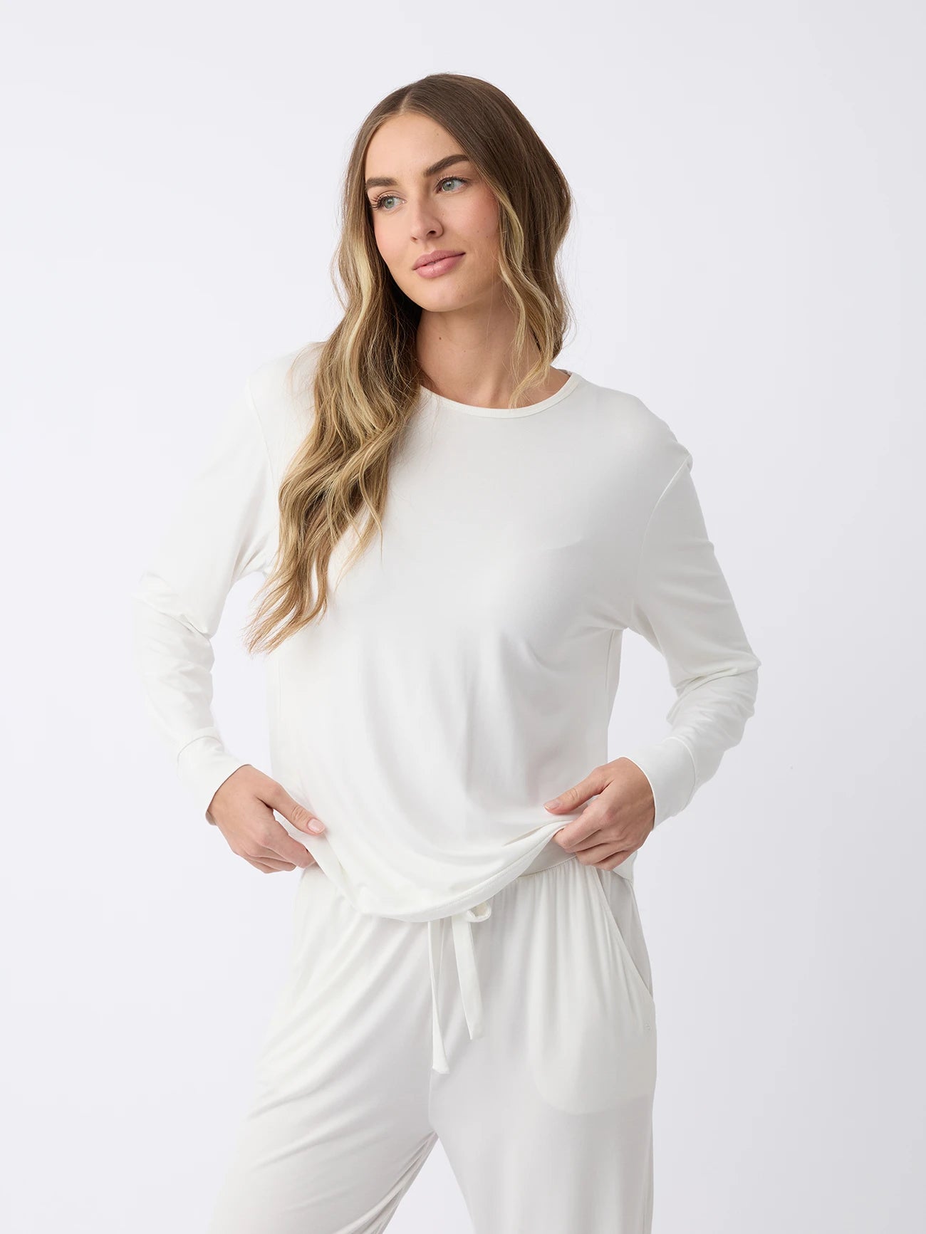 A woman in a Women's Stretch Knit Bamboo Long Sleeve Lounge Tee by Cozy Earth stands against a plain white background. Their long, wavy hair frames a relaxed expression as they look to the side, holding the fabric of the top with one hand. 