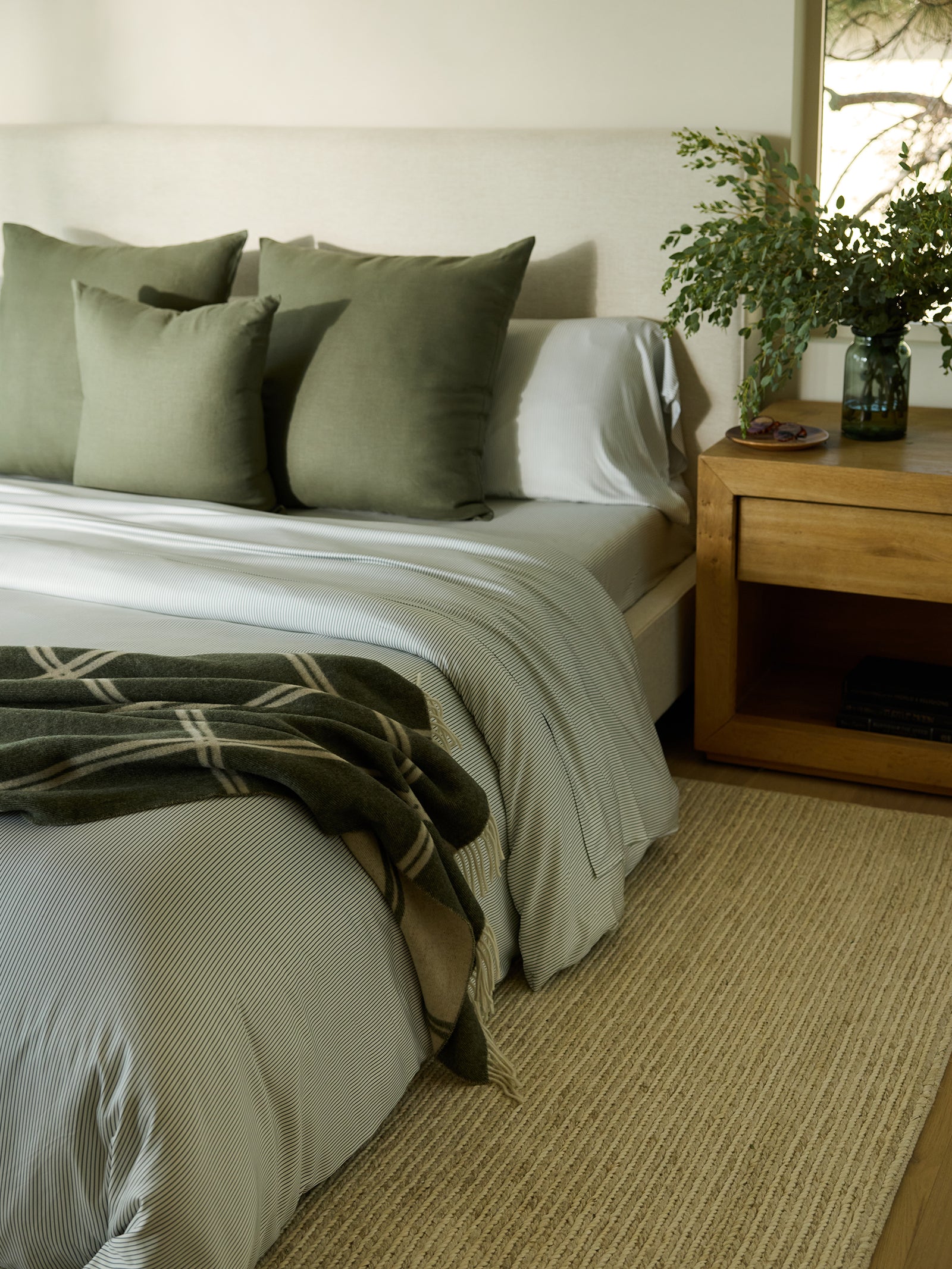 A cozy bedroom with a neatly made bed featuring a Bamboo Duvet Cover by Cozy Earth, accented with olive green pillows and a plaid throw. A wooden nightstand holds a vase with greenery. Soft natural light filters in, illuminating the beige and green decor. 