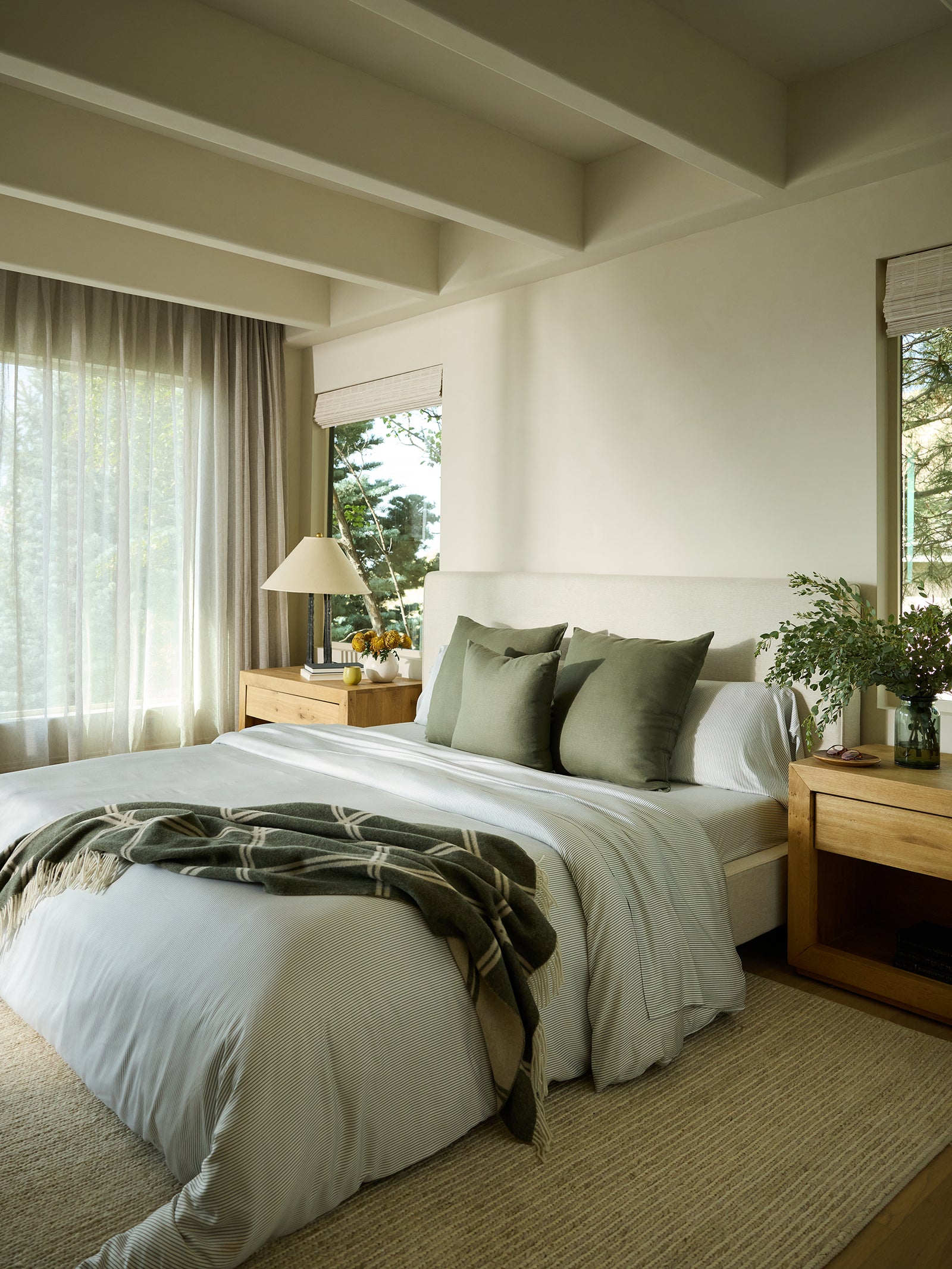 A cozy bedroom with a neatly made bed showcasing white Bamboo Flat Sheets from Cozy Earth and green pillows. A green throw is draped over the bed. Wooden nightstands hold a lamp and a vase with greenery, while sunlight filters through sheer curtains next to large windows. 