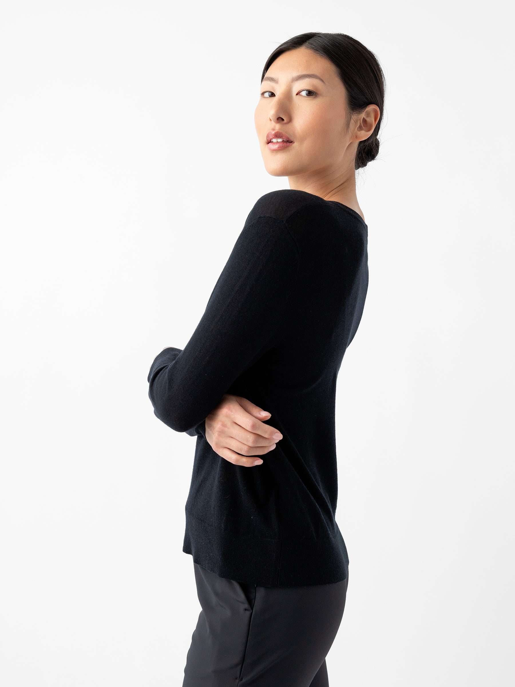 A woman with dark hair tied back wears the Women's AirKnit V-Neck Sweater by Cozy Earth, paired with black pants. She stands sideways, looking over her shoulder towards the camera against a plain white background. |Color:Jet Black