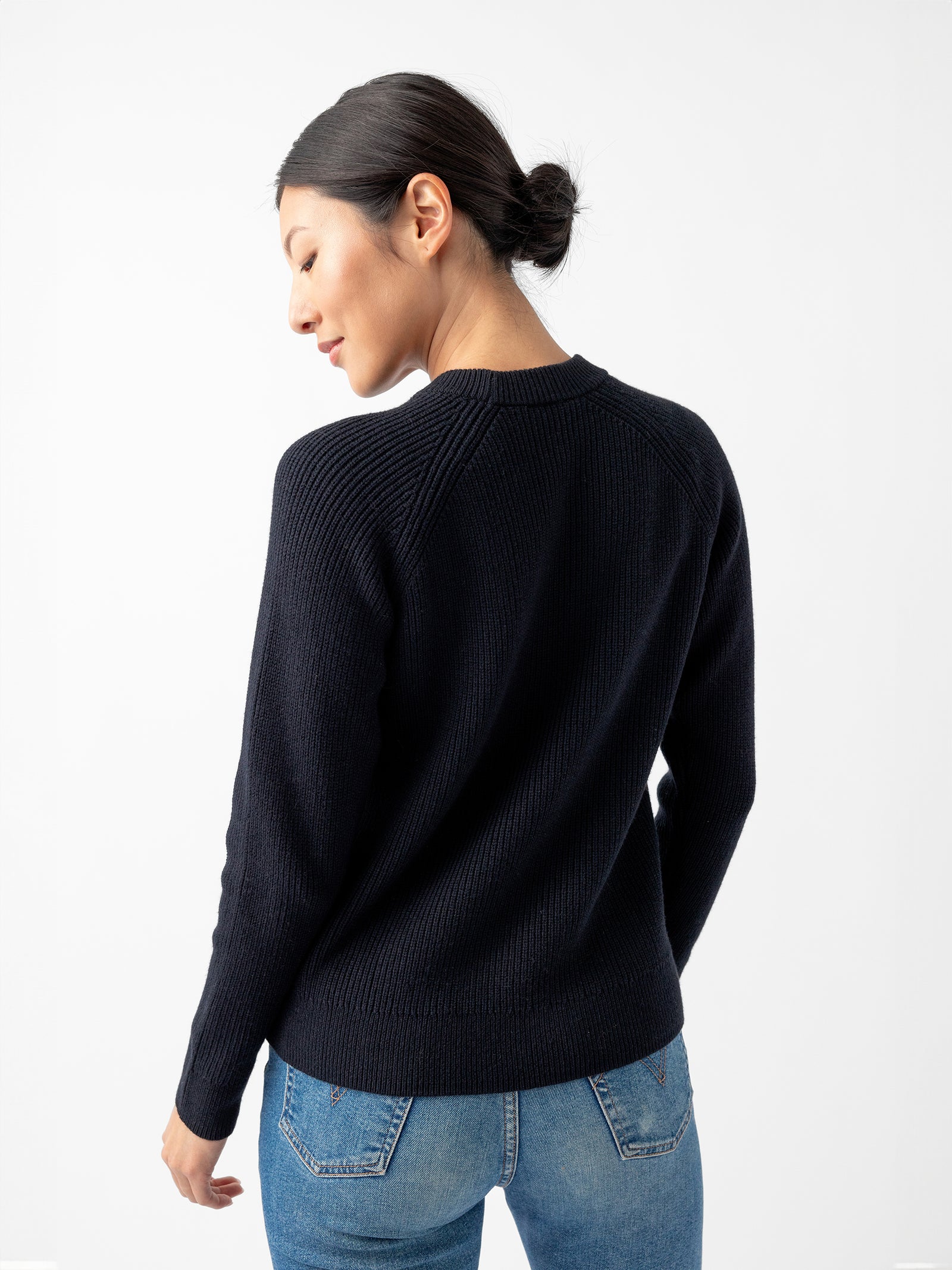 A person with dark hair styled in a low bun is wearing the Women's Classic Crewneck by Cozy Earth and blue jeans, standing with their back facing the camera. The background is white and plain. 