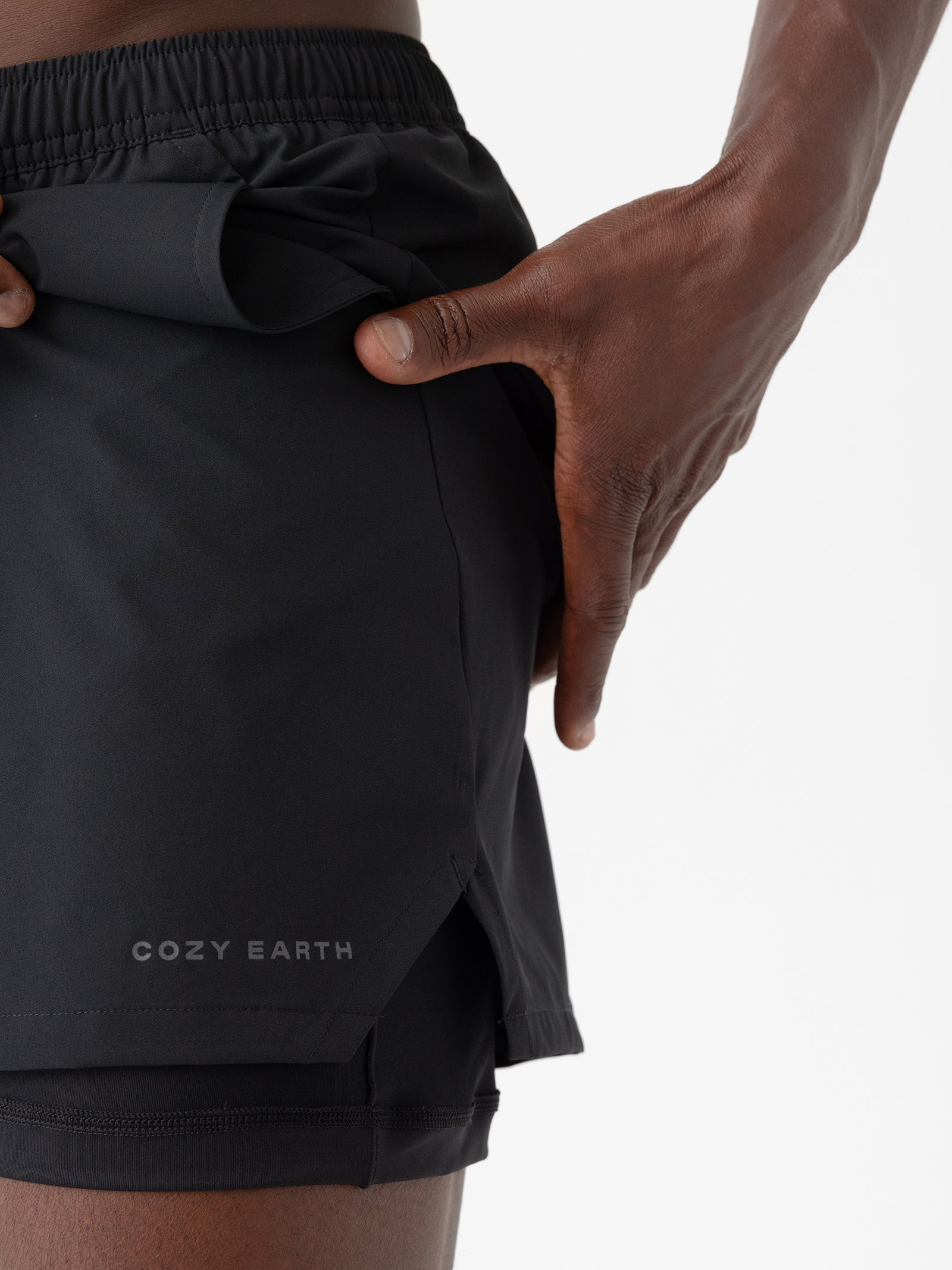 A close-up shows a person wearing Cozy Earth's Men's Performance Sleep Short in black, featuring a small slit at the hem and the brand name "Cozy Earth" printed near the edge. The person is adjusting the shorts with one hand against a plain white background. 