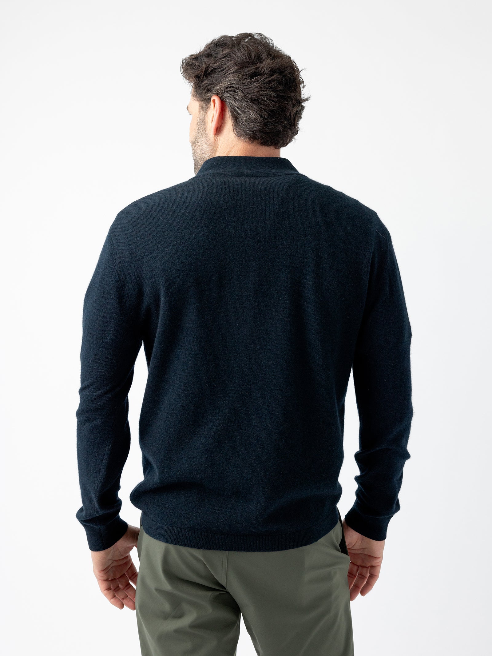 A man with short dark hair is seen from the back, wearing a long-sleeved, dark blue Men's Quarter Zip Sweater by Cozy Earth and olive green pants. He stands against a plain white background. 