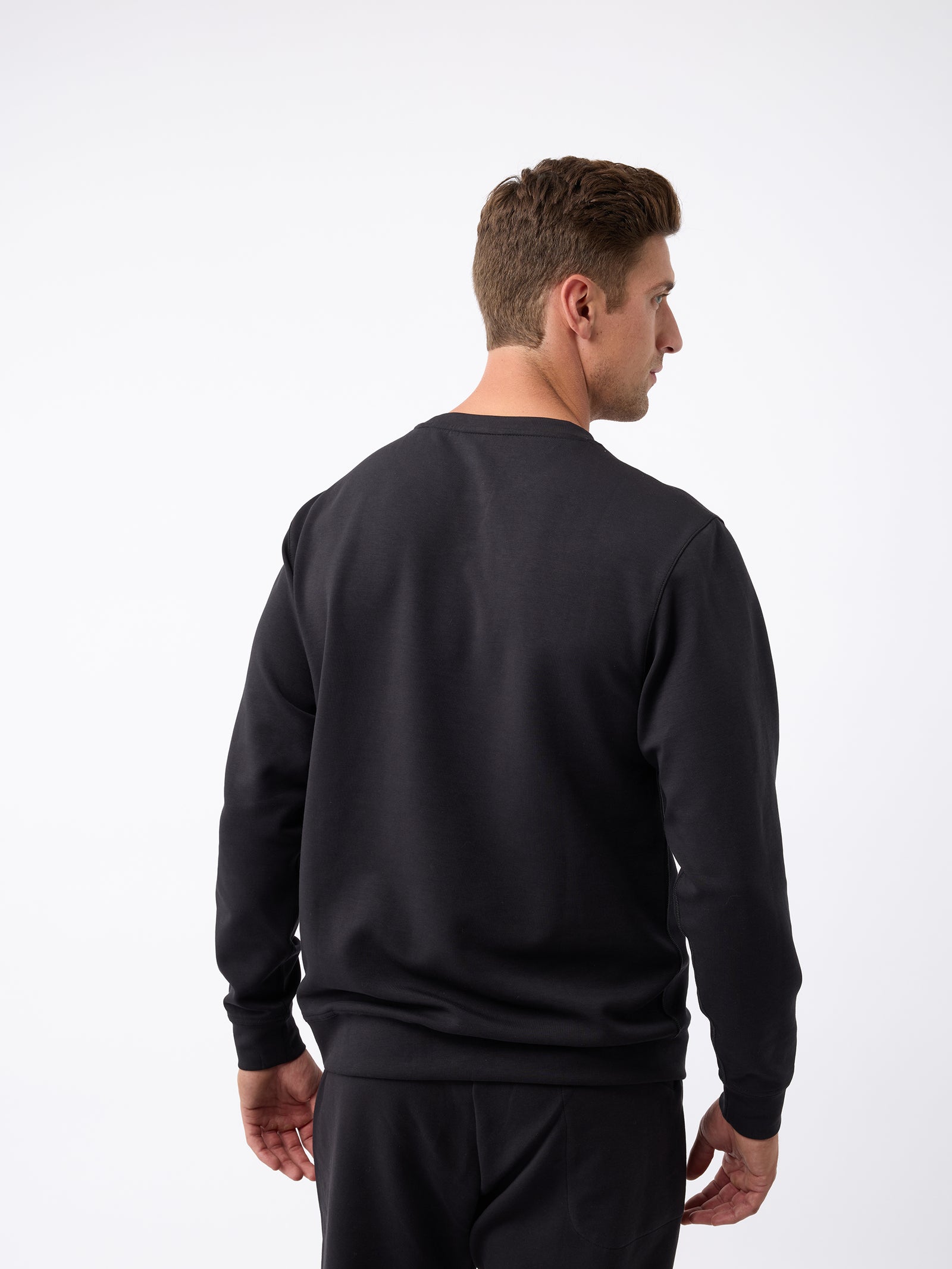 A man wearing a Cozy Earth Men's StretchTech Crewneck in black, along with matching pants, is seen from the back looking to his left. He has short brown hair and is standing against a plain white background. 