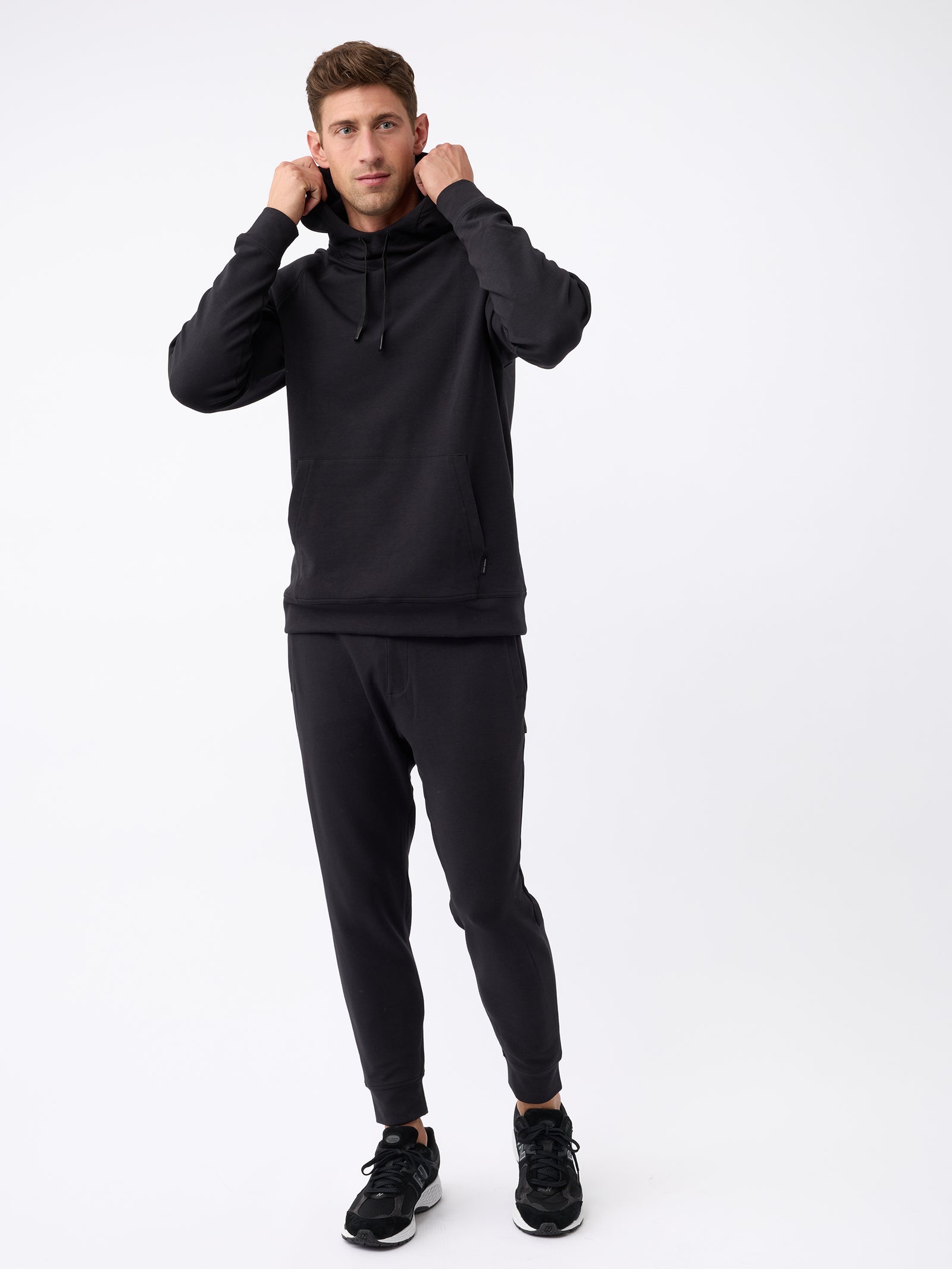 A person with short brown hair is shown posing against a plain white background. They are adjusting the hood of their Cozy Earth Men's StretchTech Hoodie and wearing matching black jogger pants and black sneakers. 