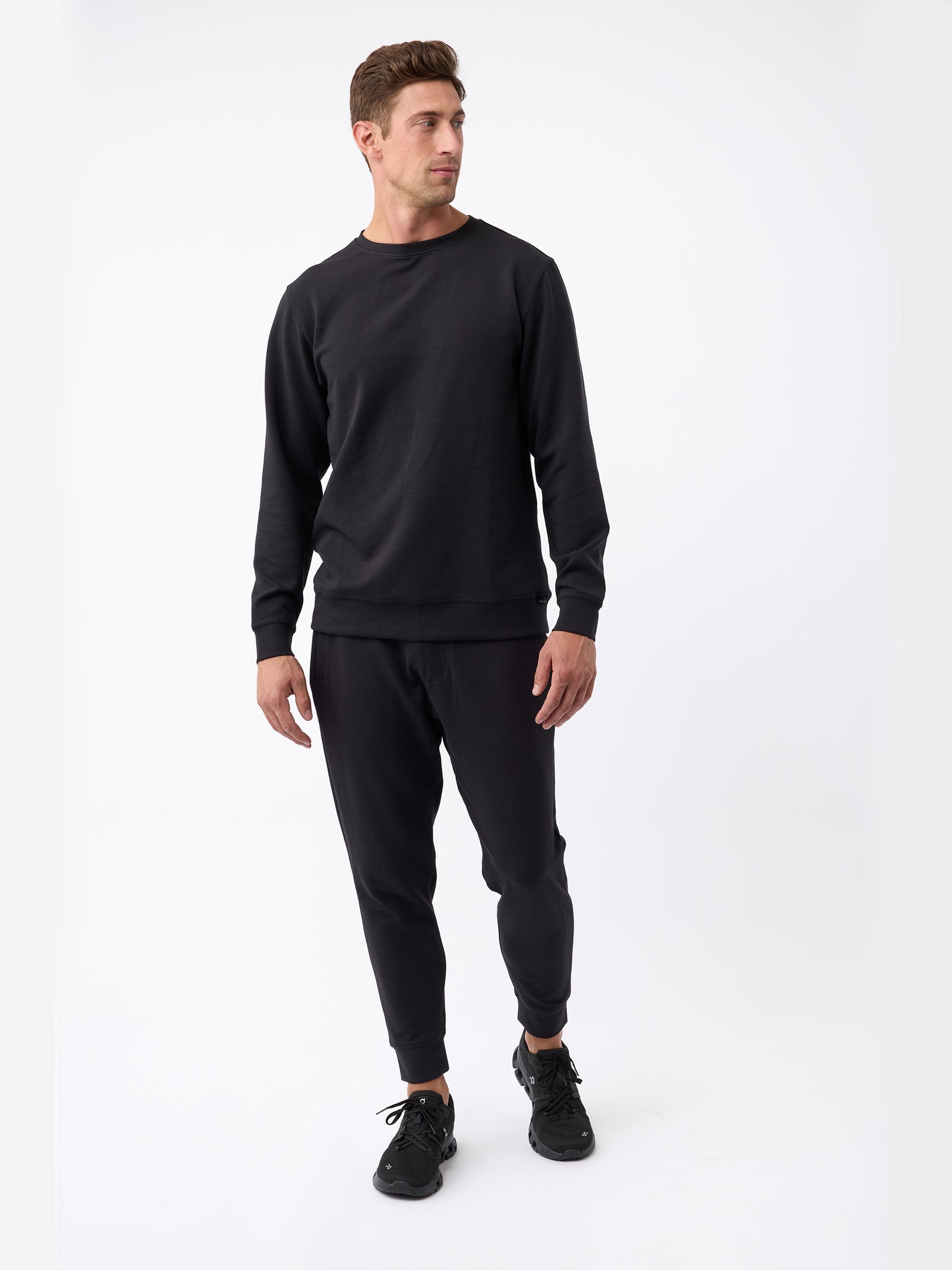 A person is standing against a plain white background, looking to the side. They are wearing a black sweatshirt, Cozy Earth's Men's StretchTech Jogger in black, and black sneakers. Their hair is short and styled, and their hands are relaxed at their sides. 