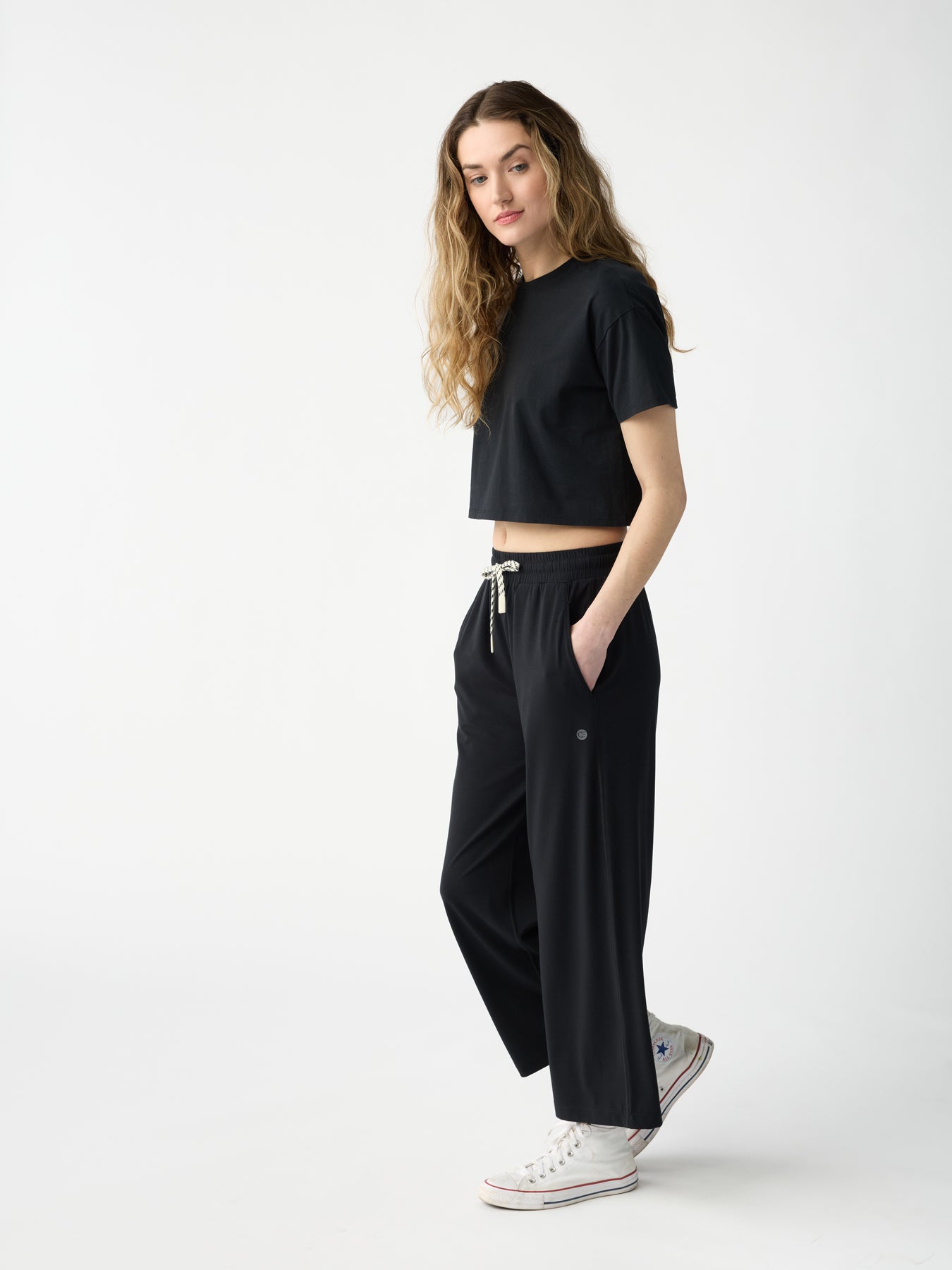 A person with long wavy hair wears Cozy Earth's Women's Studio Cropped Wide Leg Pant in black and a black crop top, paired with white sneakers. They stand sideways in a minimalistic setting against a light background. |Color:Jet Black