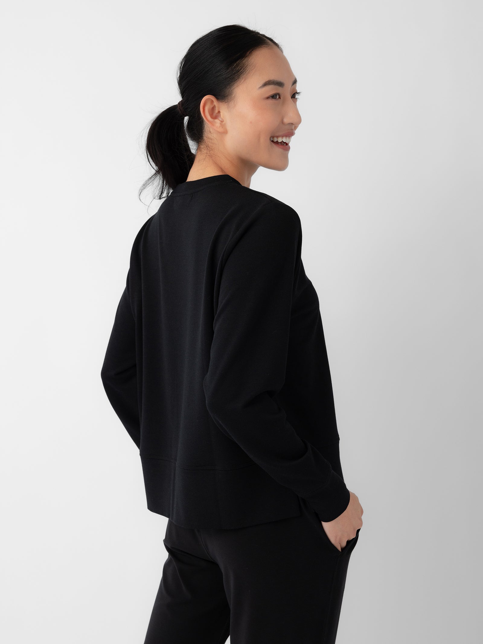 A woman with black hair tied in a ponytail is smiling and looking to the side. She is wearing the Women's StudioLite Crewneck by Cozy Earth and black pants, standing against a plain white background. 