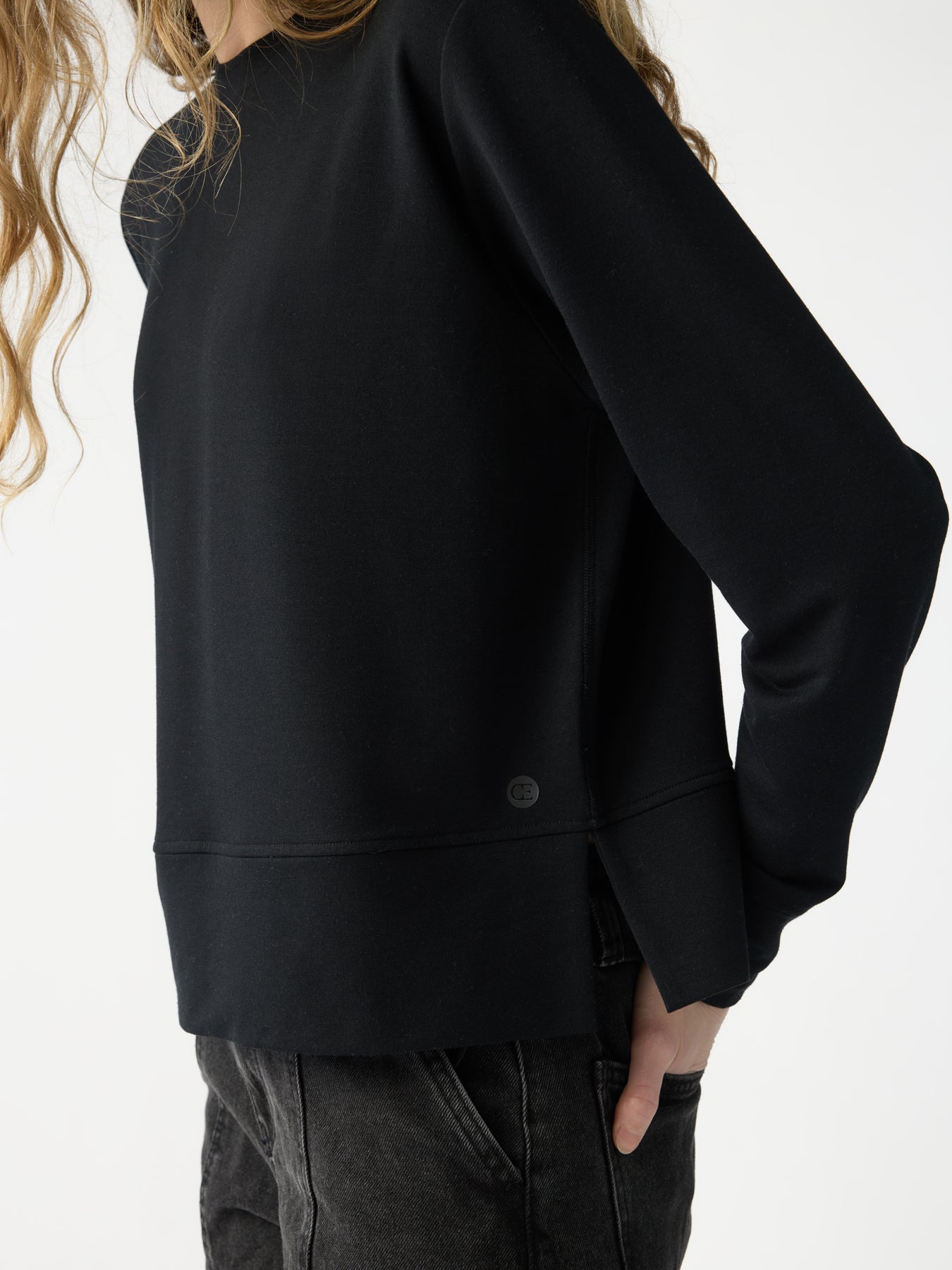 A person with long, wavy hair wearing a loose-fitting Women's StudioLite Crewneck by Cozy Earth and dark jeans stands sideways with one hand in their pocket on a plain background. |Color:Jet Black