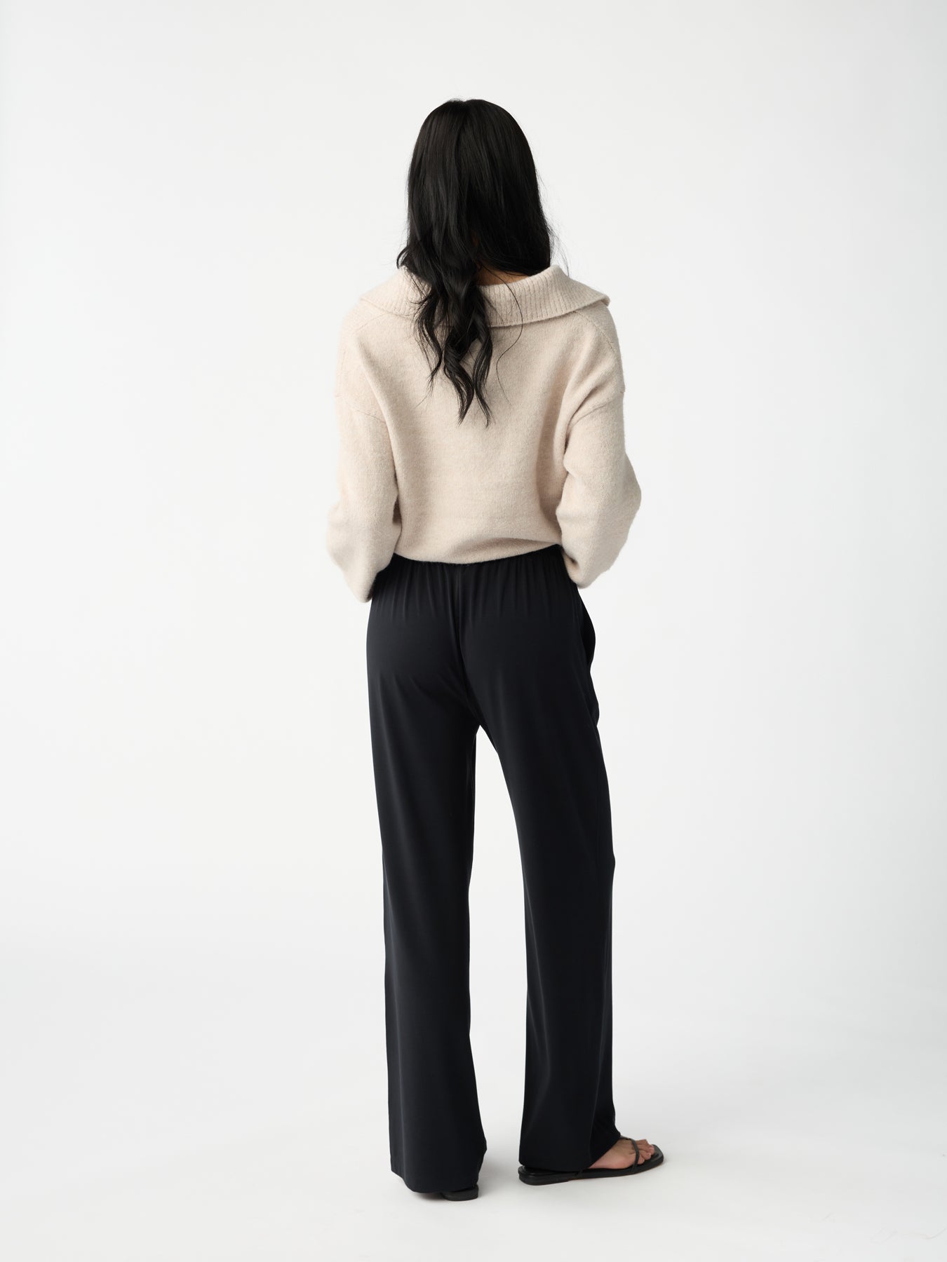 A person with long black hair, facing away from the camera, wears a cream sweater and Cozy Earth's Women's Studio Wide Leg Pant against a plain white backdrop. 