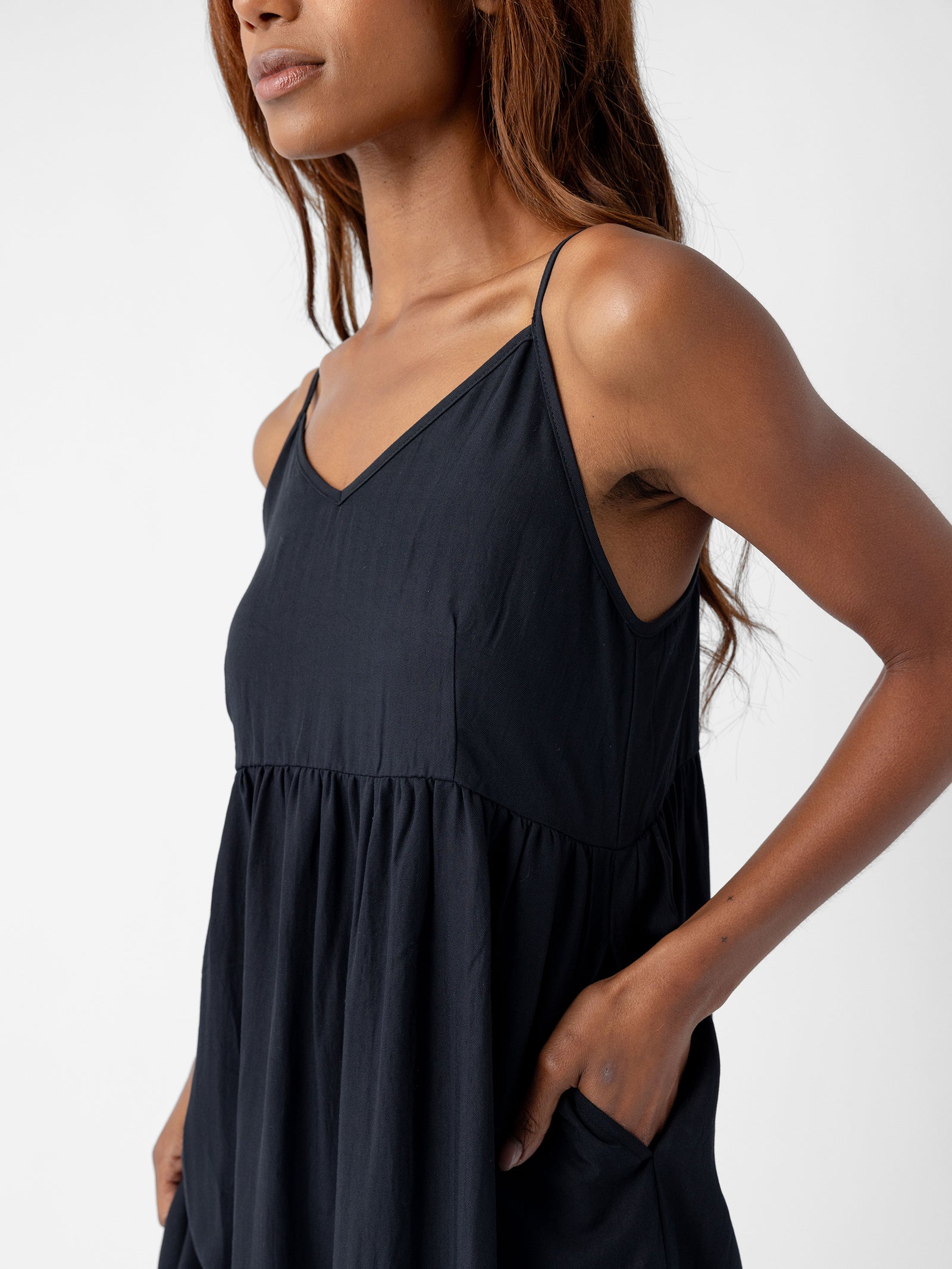 A person with long, brown hair is wearing the Women's Sunset Gathered Dress by Cozy Earth, a black, sleeveless, V-neck dress with thin straps. The dress features a flowy design with a high waist. The person's right hand is casually placed in their pocket, and the background is plain white. 