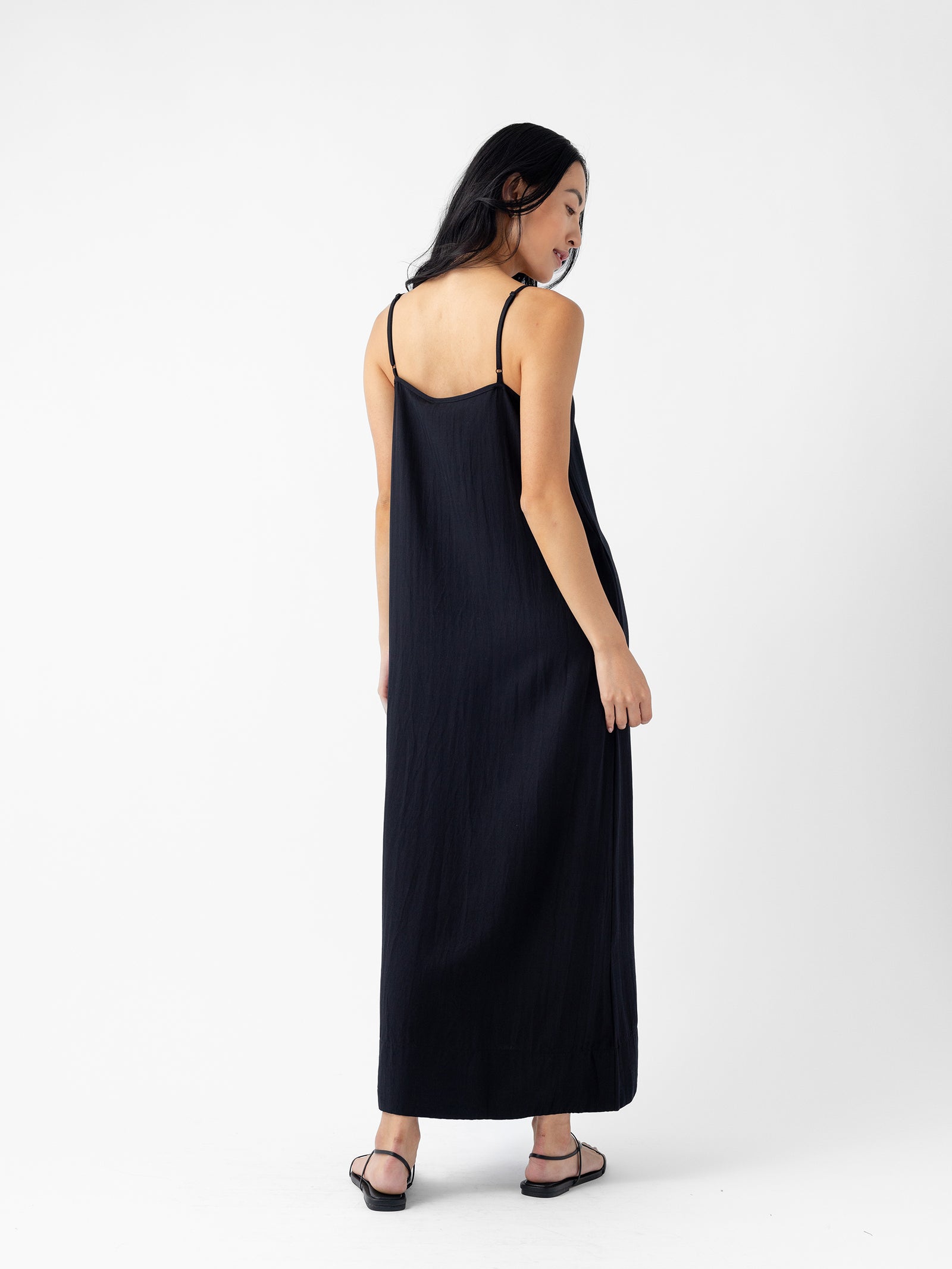 A woman with long black hair wearing the Cozy Earth Women's Sunset Slip Dress and black sandals stands facing away, looking over her right shoulder against a plain white background. 