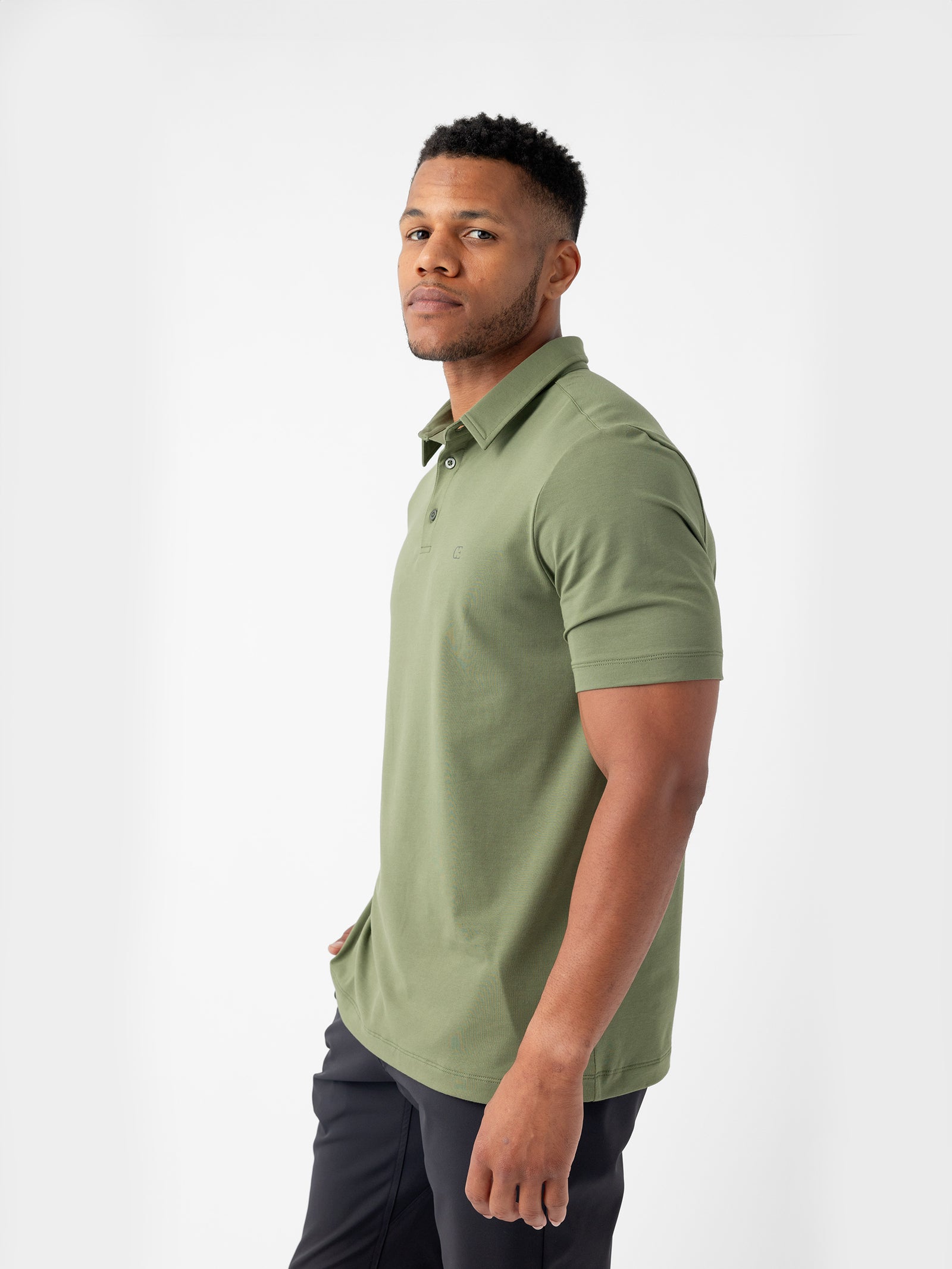 A man dressed in a Men's Everyday Polo by Cozy Earth in light green and dark pants is standing and looking at the camera against a plain white background. 