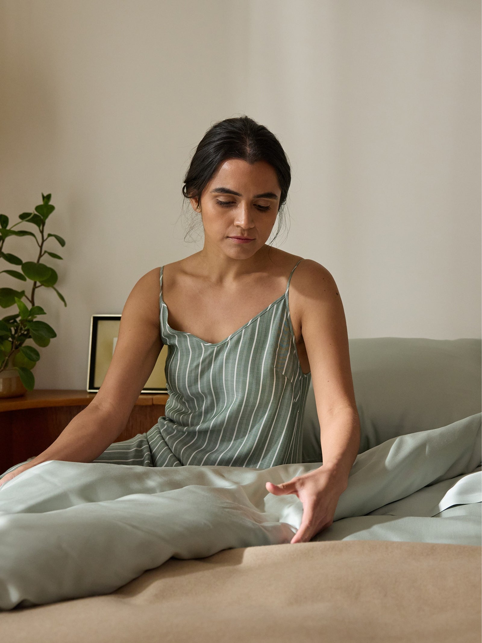 A person sits on a bed with Cozy Earth's Bamboo Sheet Set, wearing a striped dress. A small plant and picture frame adorn the wooden nightstand behind them in the softly lit room. 