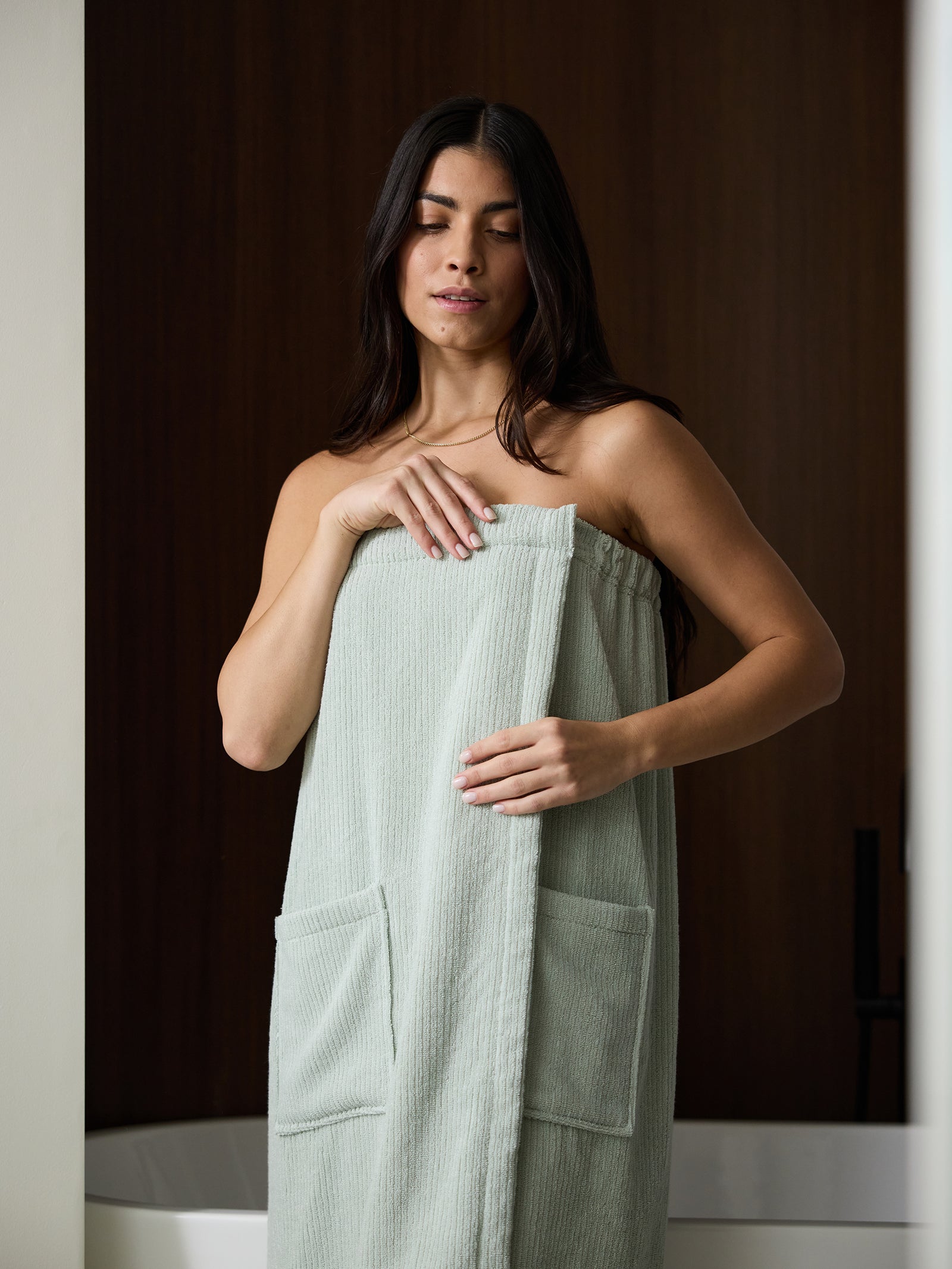 A person with long dark hair stands in a room, wrapped in a Cozy Earth Ribbed Terry Bath Wrap with pockets. The individual’s eyes are closed, and one hand is adjusting the wrap at their chest. The background features a dark wood panel and a white bathtub. 