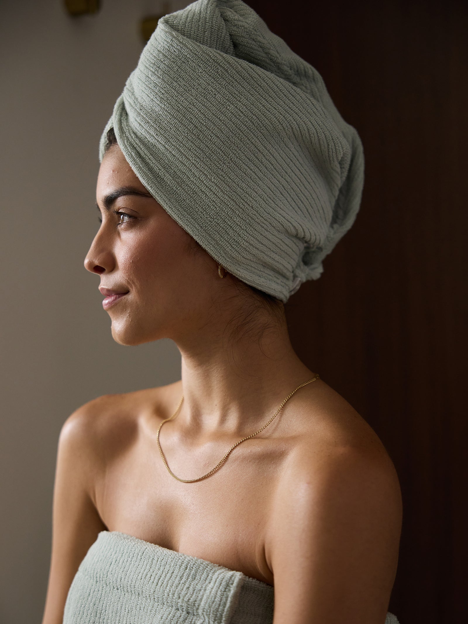 A woman with a serene expression is wrapped in Cozy Earth's textured towel, the Ribbed Terry Hair Towel, and her hair is wrapped in a matching turban. She is wearing a delicate gold necklace and stands against a neutral background, illuminated by soft lighting. 