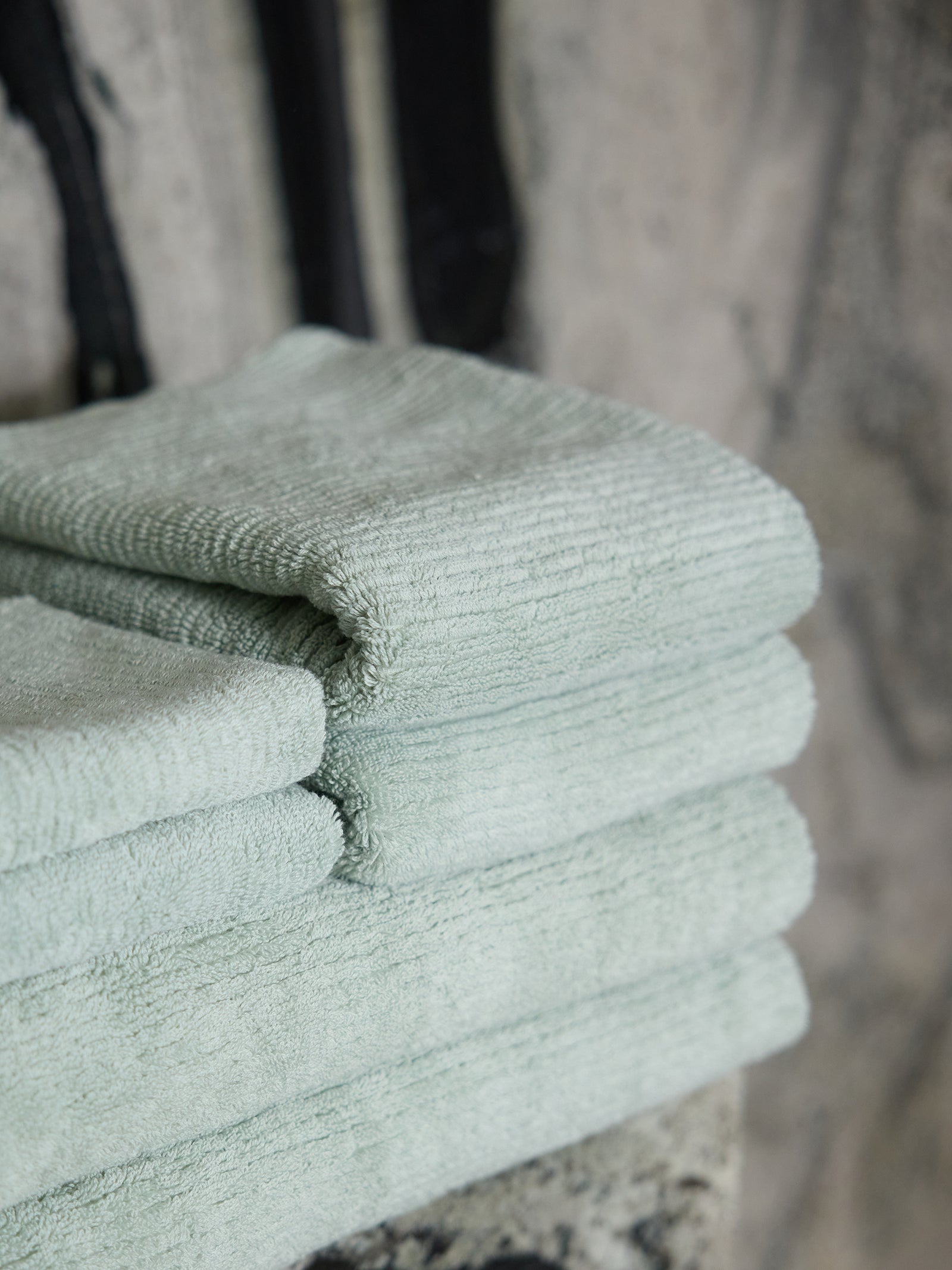 A stack of neatly folded Ribbed Terry Hand Towels from Cozy Earth, in a light green hue, is placed on a surface against a blurry, neutral-colored background. The towels have a soft, plush texture, indicating they are made of absorbent material. 