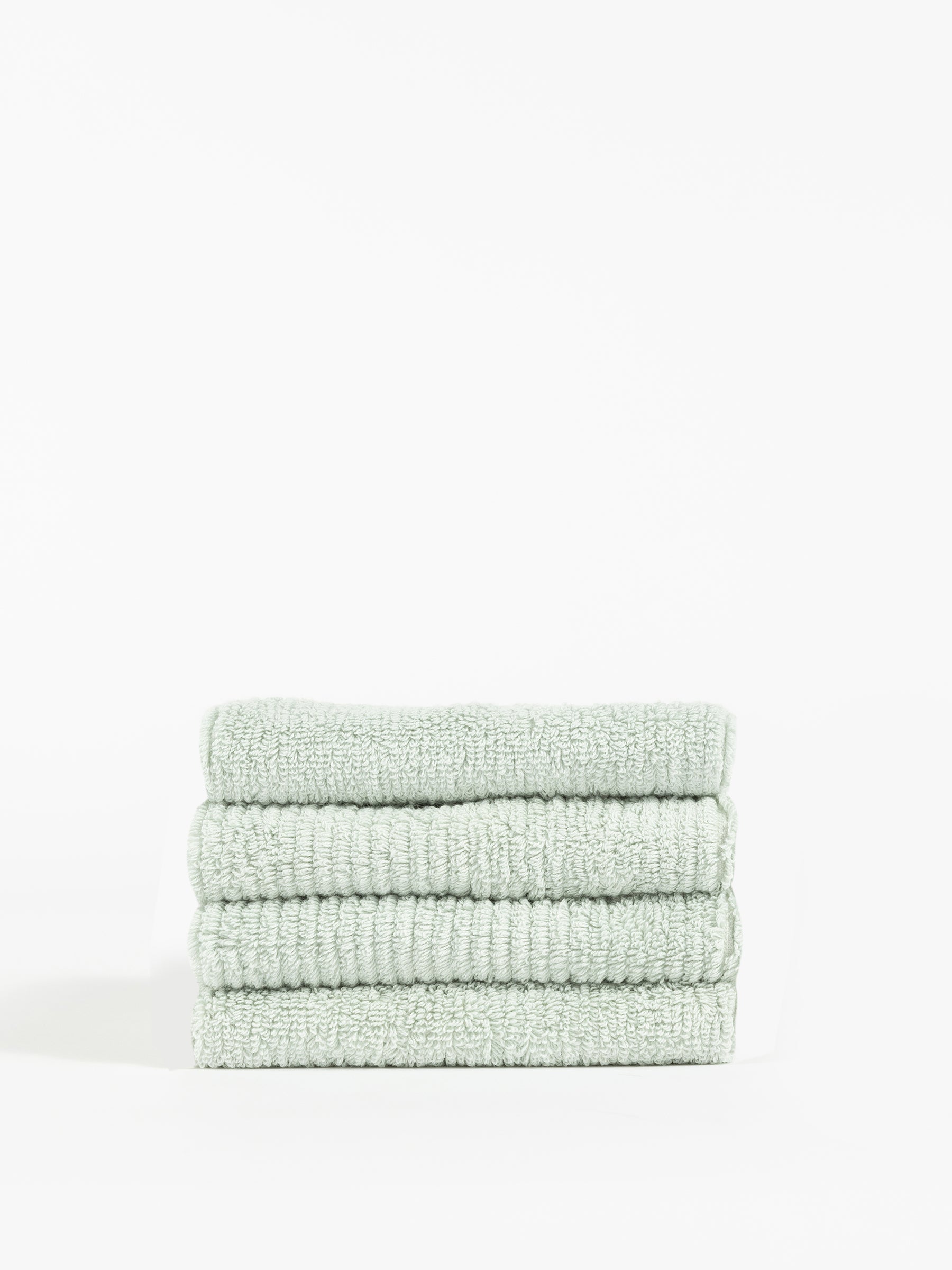 A stack of four neatly folded, Cozy Earth Ribbed Terry Washcloths in light green is placed against a plain white background. The texture of these washcloths appears soft and plush, with visible ribbed detailing in the fabric. The overall look is clean and minimalistic. |Color:Laurel