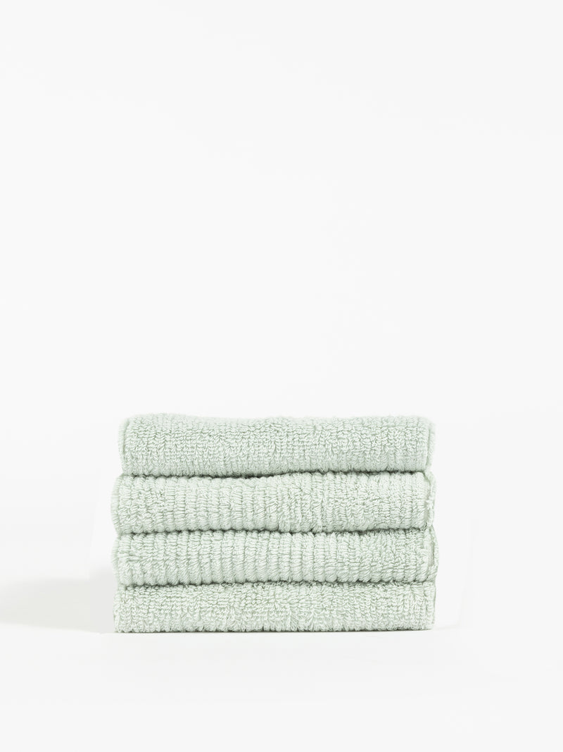 A stack of four neatly folded, Cozy Earth Ribbed Terry Washcloths in light green is placed against a plain white background. The texture of these washcloths appears soft and plush, with visible ribbed detailing in the fabric. The overall look is clean and minimalistic.