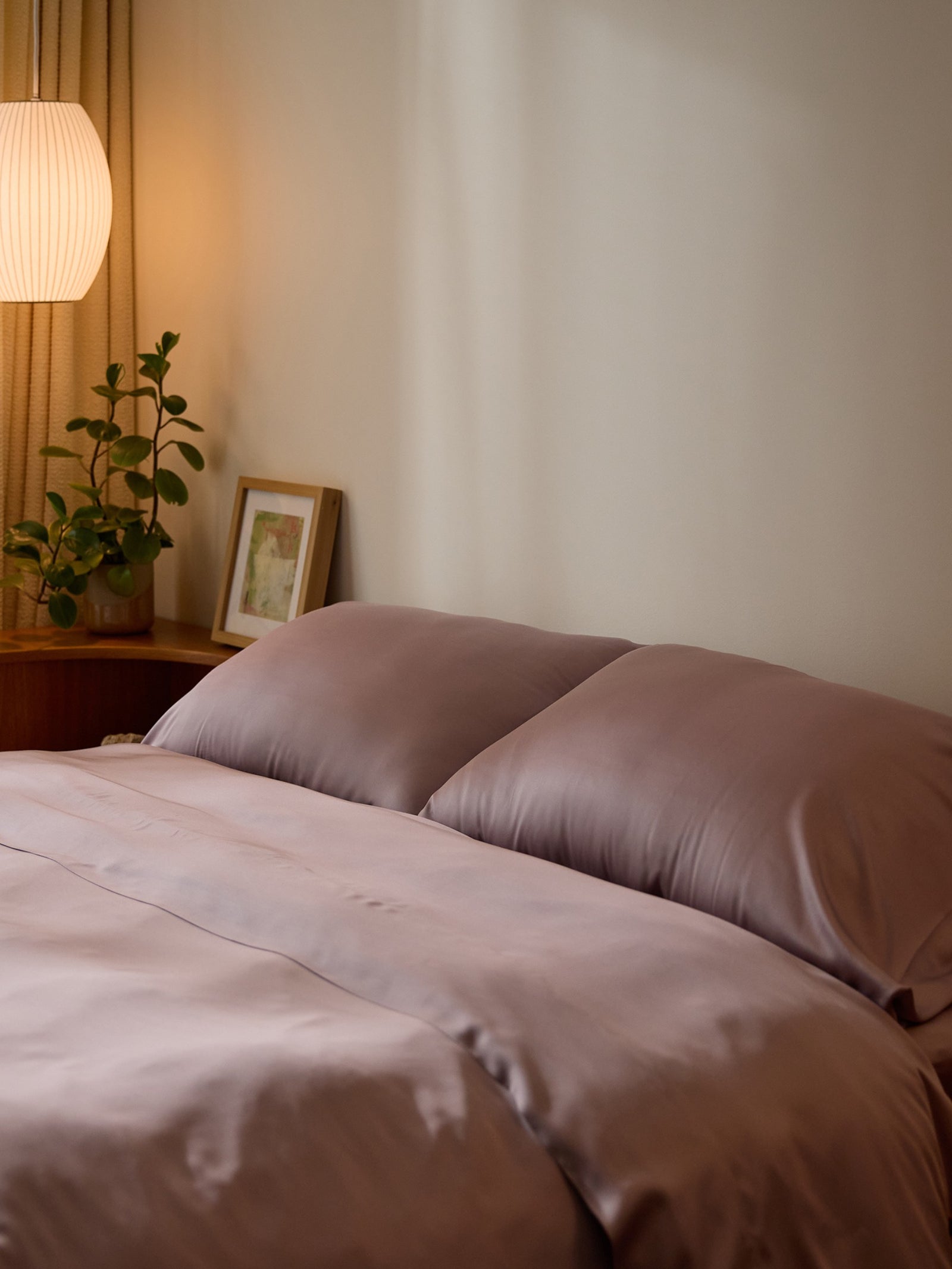 A cozy bedroom scene with a neatly made bed featuring the Bamboo Duvet Cover by Cozy Earth in lavender, accompanied by pillows. A small framed picture and potted plant sit on the bedside table next to a softly glowing lamp, creating a warm, calming ambiance. 