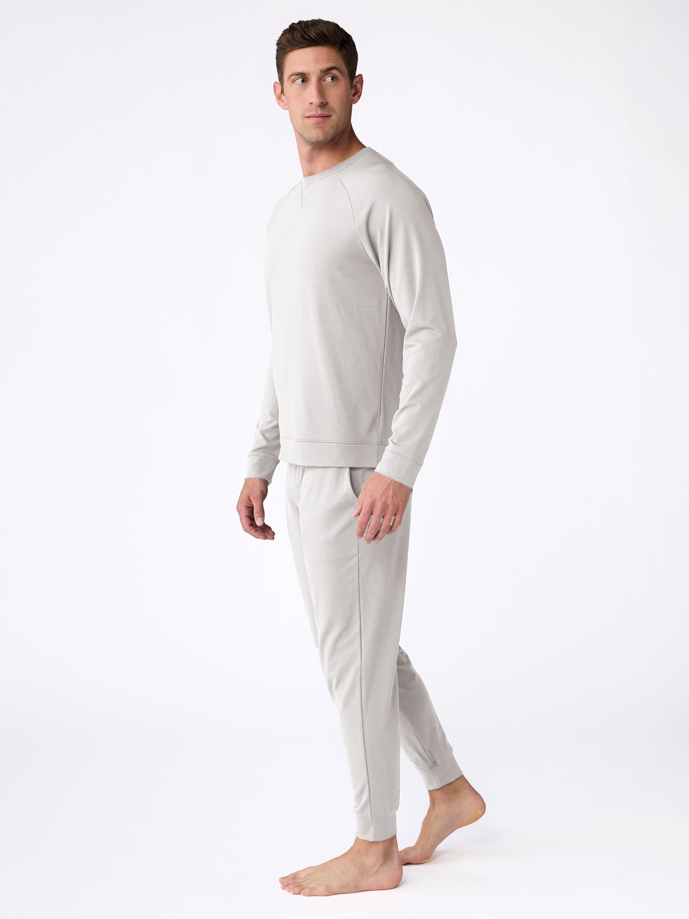 Modeling on a plain white background, an individual is barefoot, dressed in the Men's Bamboo Jogger Set from Cozy Earth. They wear a light gray long-sleeve sweatshirt and matching sweatpants while turned slightly to the side, gazing forward. 