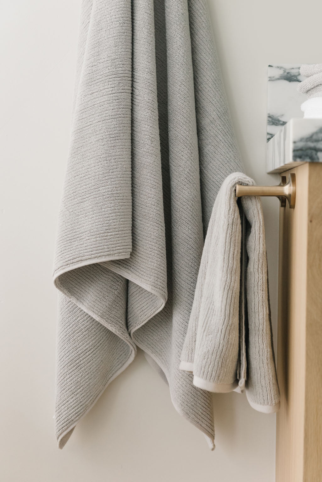 Ribbed Terry Bath Towel and Hand Towel in the color Light Grey. Photo of Ribbed Terry Bath Towel and Hand Towel taken as the Light Grey Ribbed Terry Bath Towel and Hand Towel is hanging from a towel hook in a bathroom. 