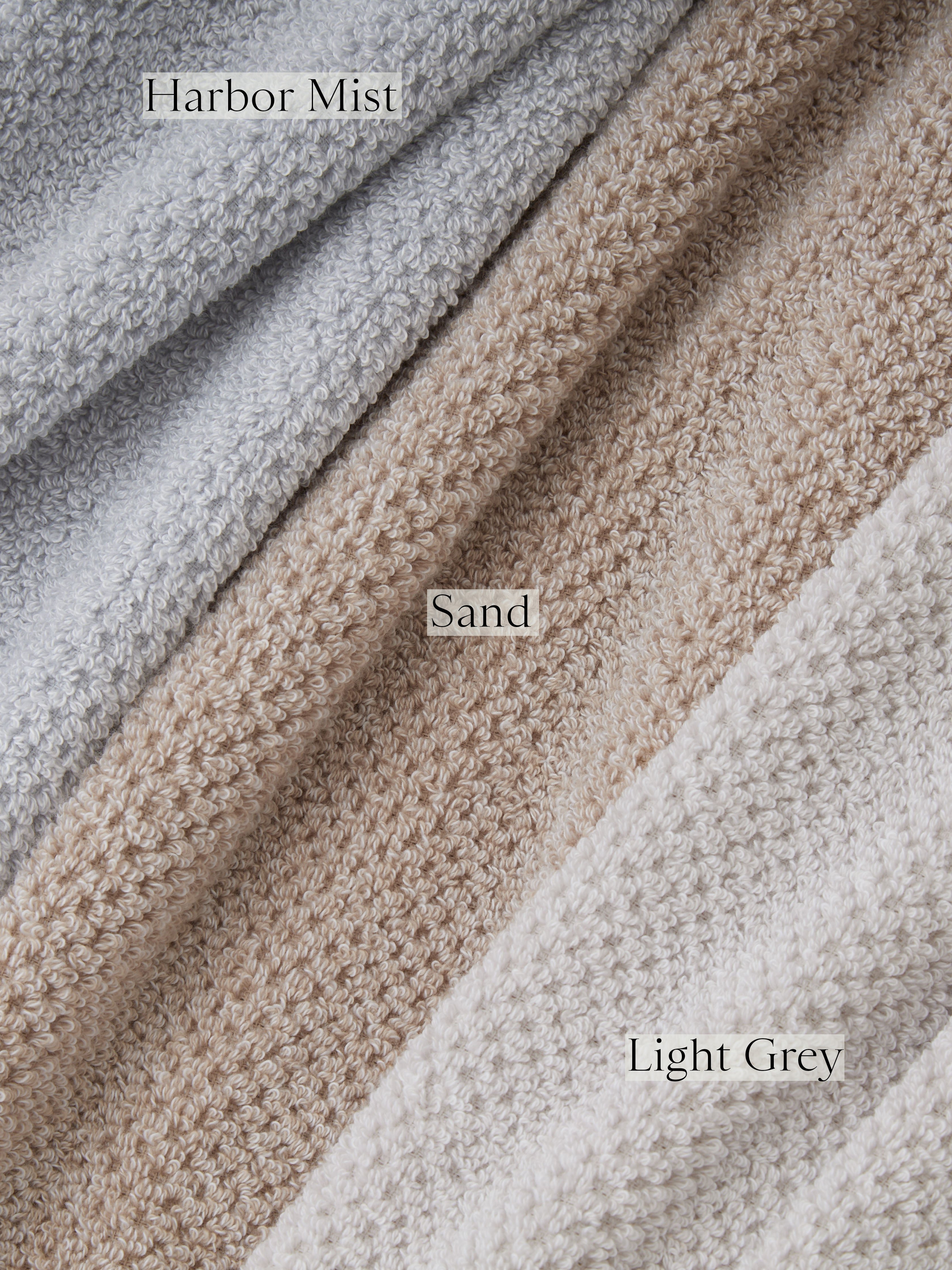 Close-up image of three textured fabric colors from the Nantucket Hand Towels collection by Cozy Earth, labeled from top to bottom: "Harbor Mist" showcasing a light gray hue, "Sand" displaying a beige hue, and "Light Grey" showing another shade of light gray. Each fabric features a subtle, intricate woven pattern.