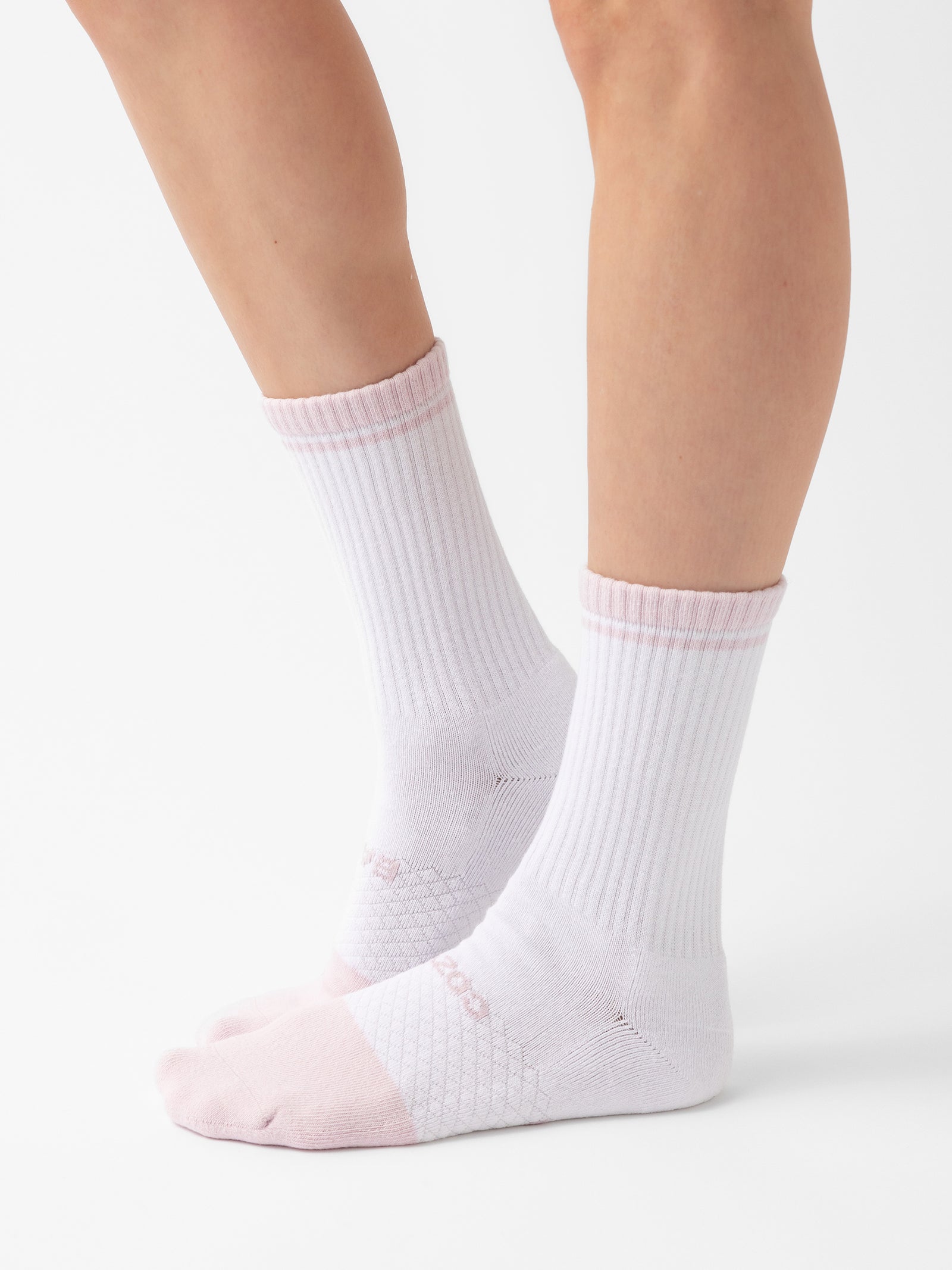 Close-up of a person wearing Essential Calf Socks by Cozy Earth in white with light pink toes and heels, and a subtle pink band at the top, standing against a plain white background. The socks feature ribbed texture above the ankle and a grid-like pattern around the arch. 