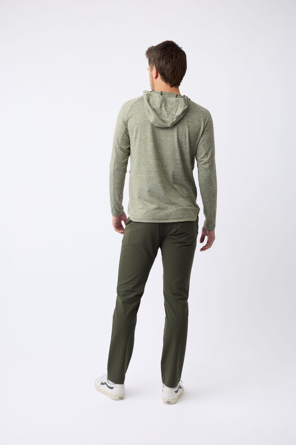 A person stands facing away from the camera against a plain white backdrop, wearing a light green Men's Lightweight Performance Hoodie by Cozy Earth, dark green pants, and white sneakers. 